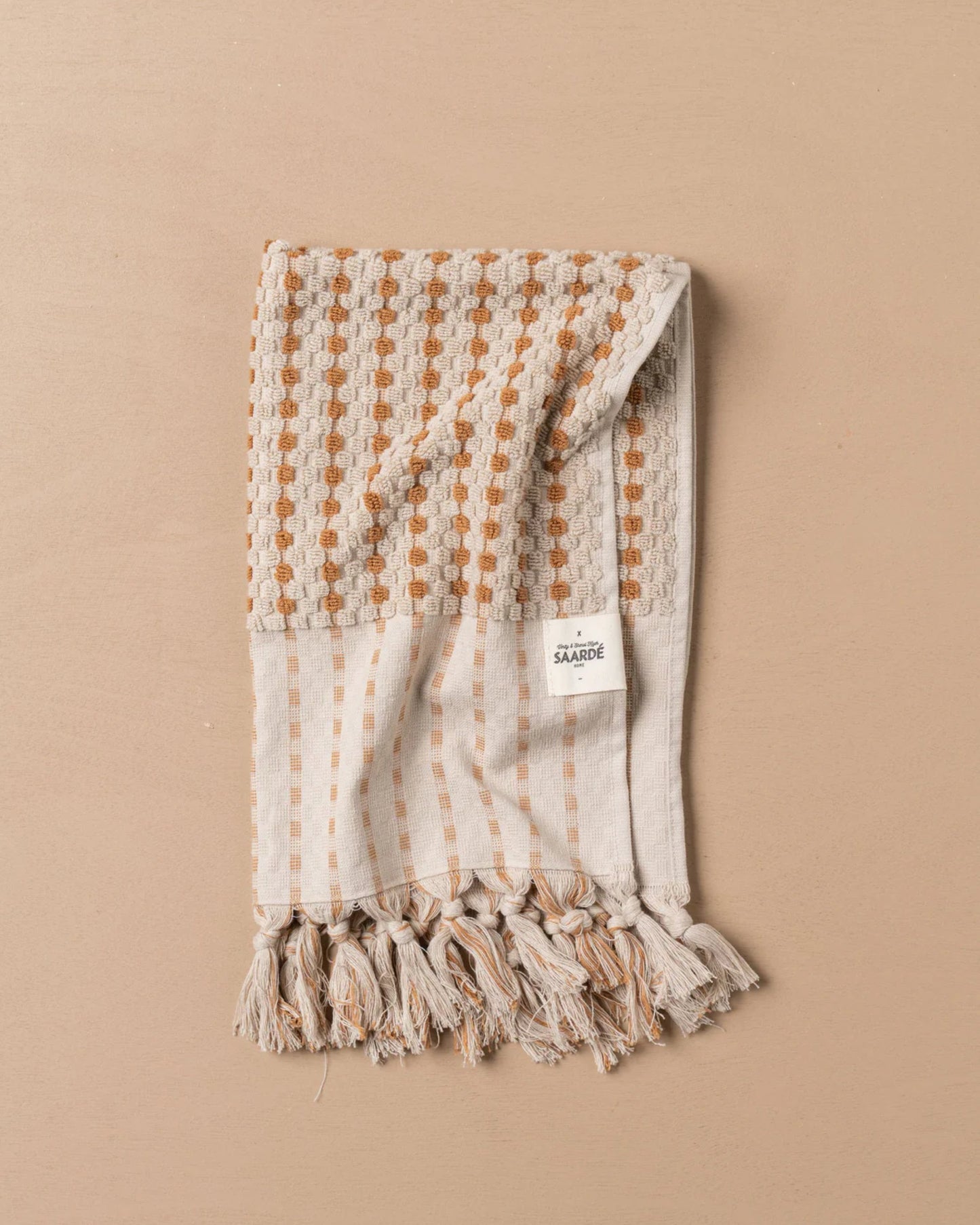 The CHICKPEA HAND TOWEL by Saarde, crafted from Turkish cotton, showcases a blend of checkered and striped designs in beige and orange. It features fringed edges and is elegantly folded on a plain beige background, with a small white label affixed to one corner.