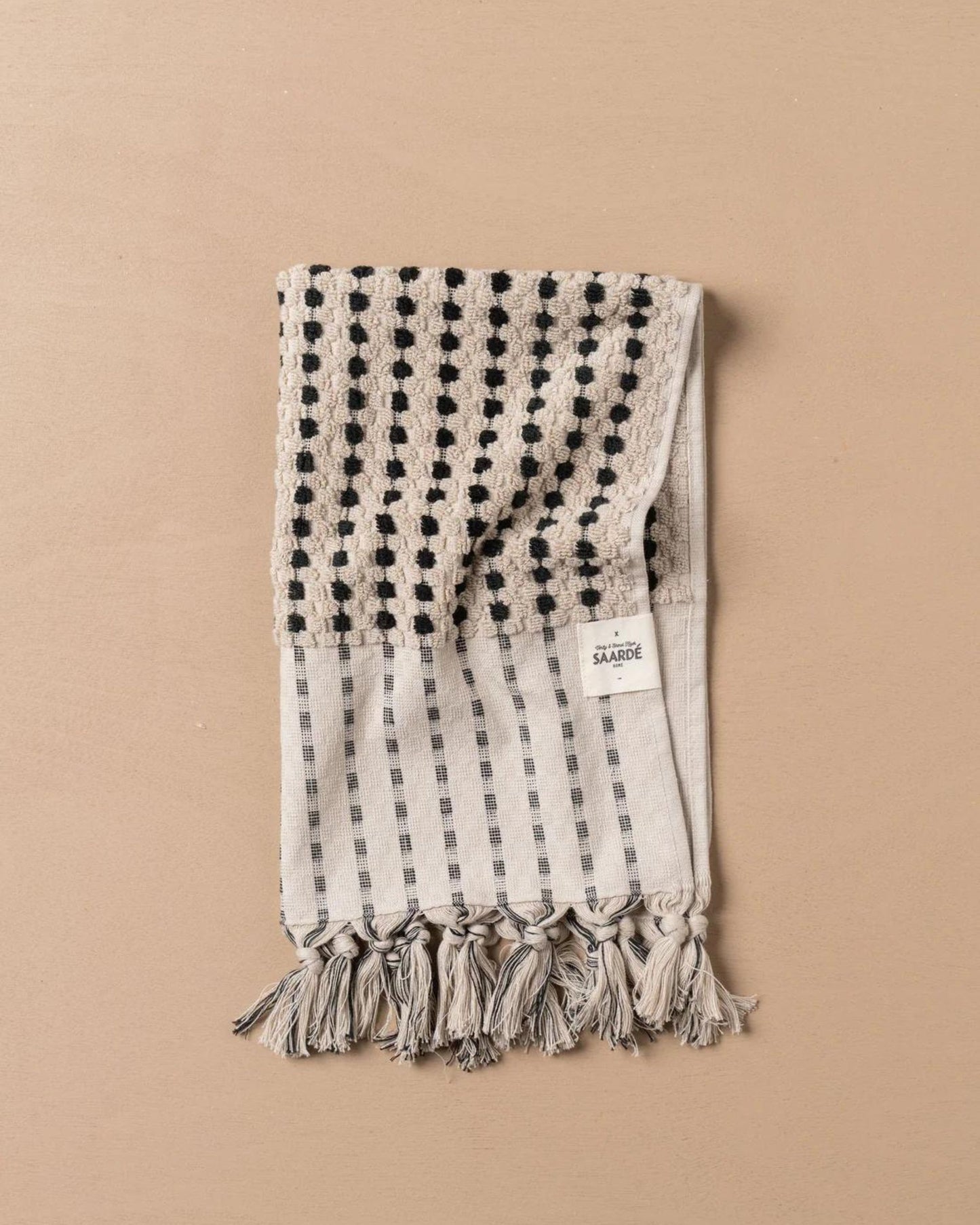 A folded CHICKPEA HAND TOWEL, designed by Saarde, showcases black geometric patterns and tassels along its edge on a plain beige background. Crafted from luxurious Turkish cotton, this beige towel is part of the esteemed Chickpea Towel Range and bears a small white tag indicating its OEKO-TEX Standard 100 certification, ensuring safety and quality.