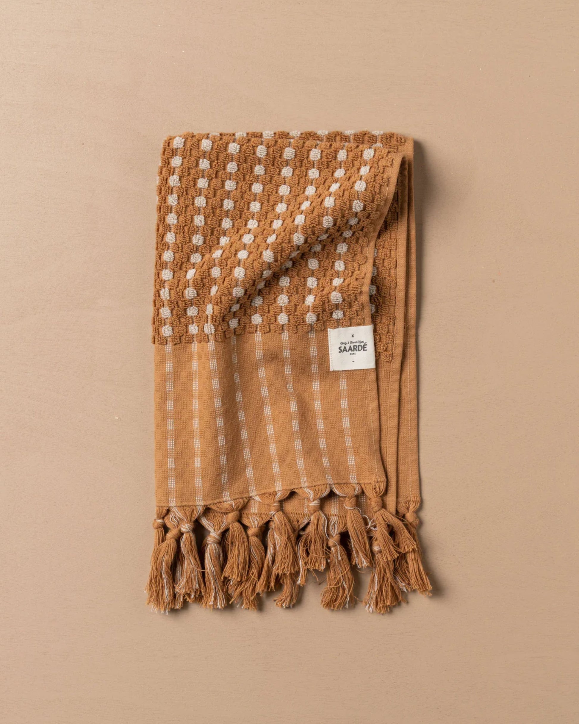 A meticulously folded textured brown scarf, made from premium Turkish cotton, adorned with white woven squares and stripes, is elegantly placed on a beige surface. It boasts tassels at one end and a small white label near the top, reflecting the quality synonymous with the Saarde CHICKPEA HAND TOWEL.