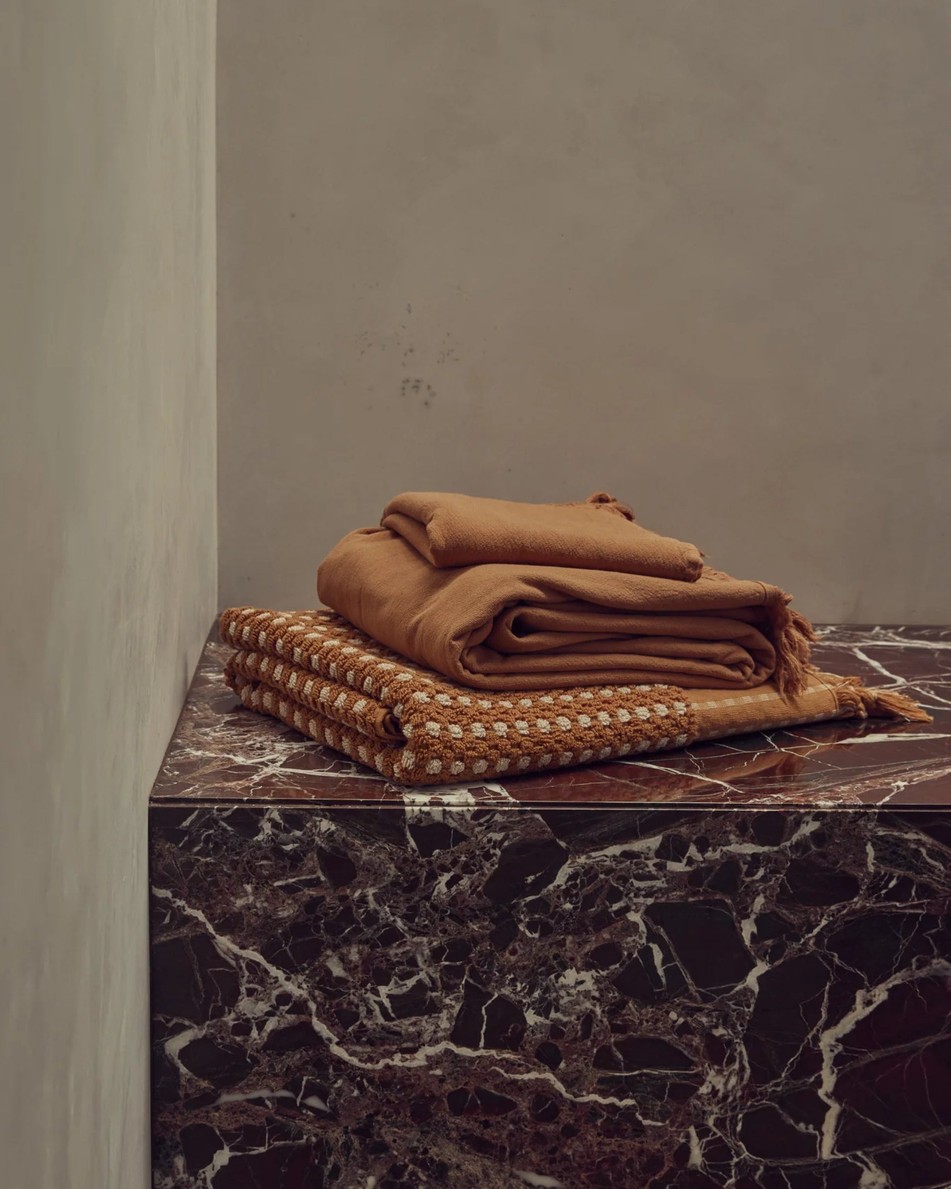 A stack of CHICKPEA HAND TOWELS by Saarde, crafted from earthy-toned luxurious Turkish cotton with elegant patterns, rests on a dark marble counter with striking white veins against a plain beige wall.