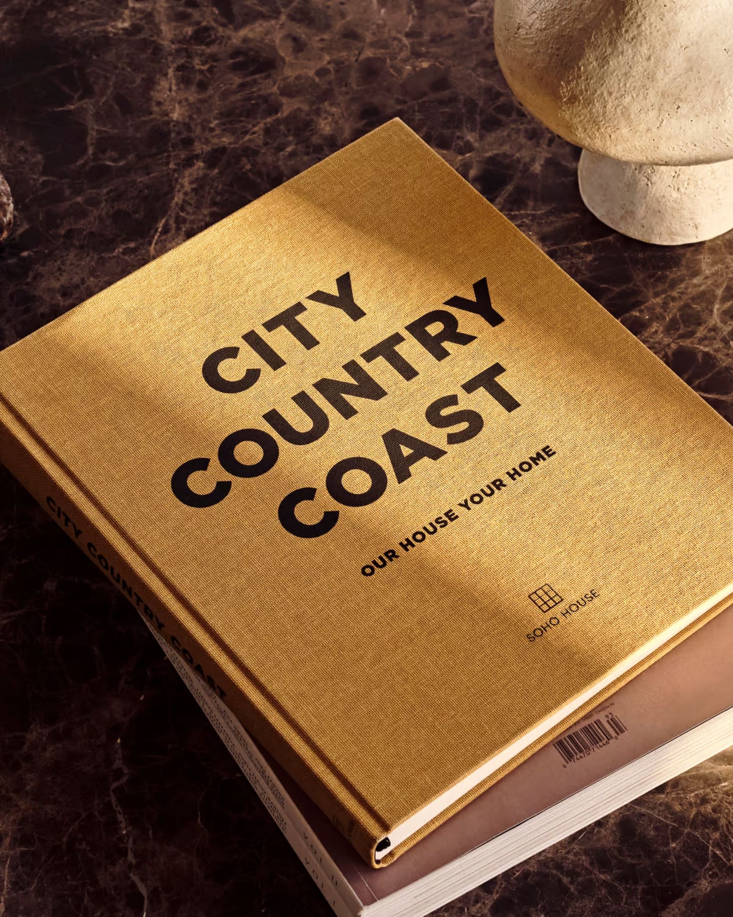 A hardcover book titled CITY COUNTRY COAST, with the brand name Bookreps elegantly inscribed beneath, rests on a dark marble surface reminiscent of Soho House's chic style, next to a round, light-colored object. A second book peeks from underneath, inviting you to discover more destinations.