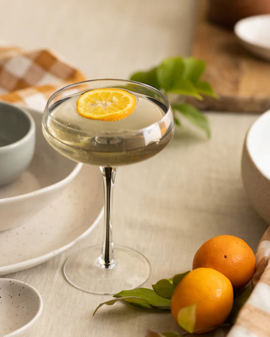 A COCKTAIL GLASS from Broste holds a drink garnished with a slice of citrus, placed on a table decorated with ceramic dishes and Nordic nature-inspired elements. Nearby, fresh oranges with leaves create an inviting ambiance enhanced by natural lighting, embodying the spirit of the Amber collection.
