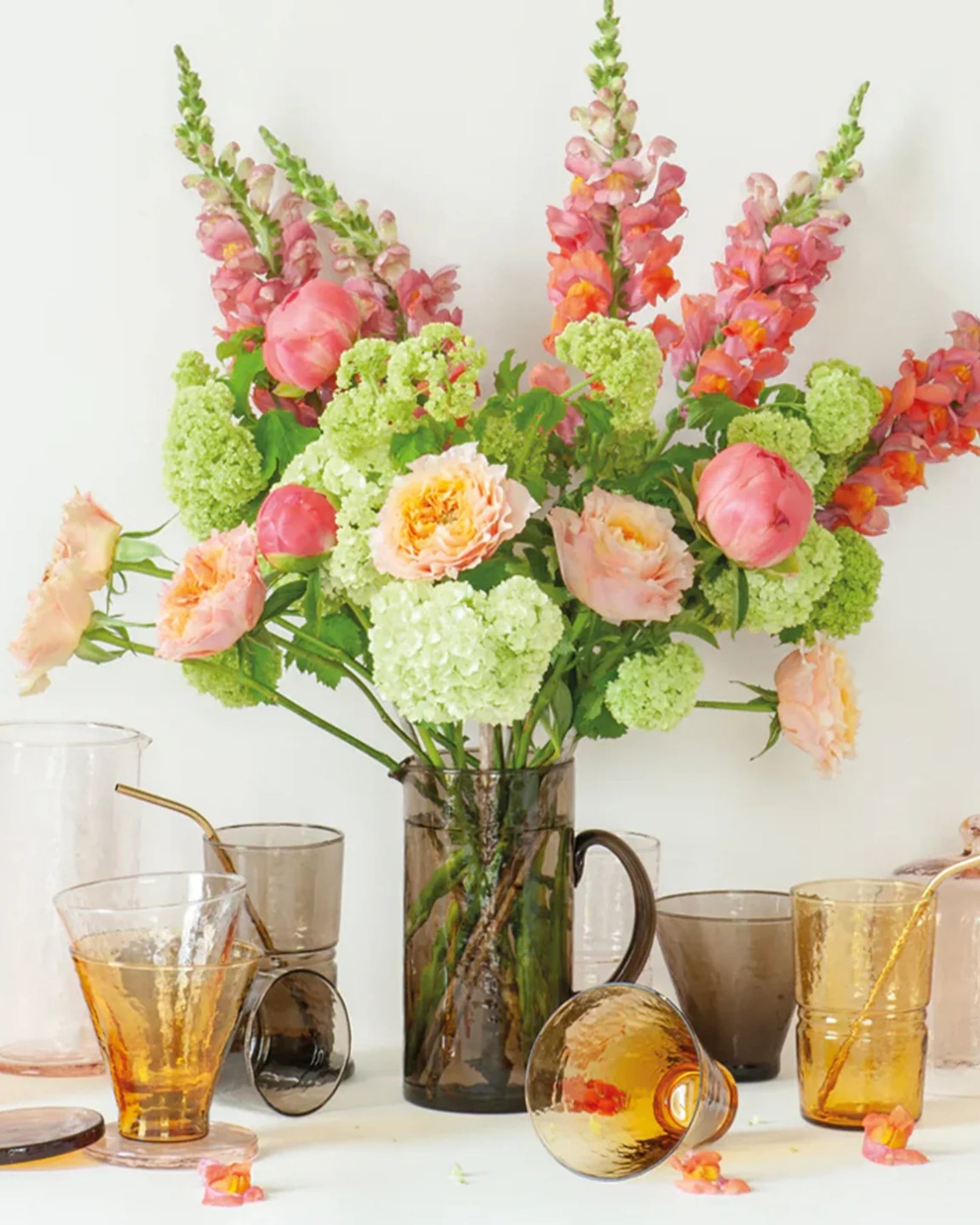 A vibrant floral arrangement with pink peonies, snapdragons, and green hydrangeas is beautifully displayed in a glass pitcher. Accompanying it is handmade glassware, including the COCKTAIL / WINE GLASS by Bianca Lorenne. The set features various colored tumblers—some paired with metal straws—and each piece showcases an organic hammered finish, all elegantly arranged on a table against a light background.