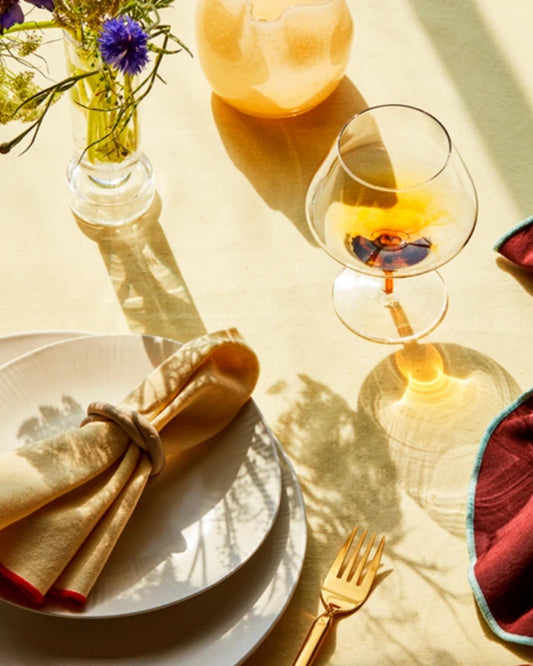 A sunlit table setting features a white plate, a beige napkin, and a gold fork. Nearby, the Broste COGNAC GLASS filled with amber liquid casts a shadow. A vase with blue flowers from the Broste Copenhagen collection and a round, textured candle add decorative touches inspired by Nordic nature.