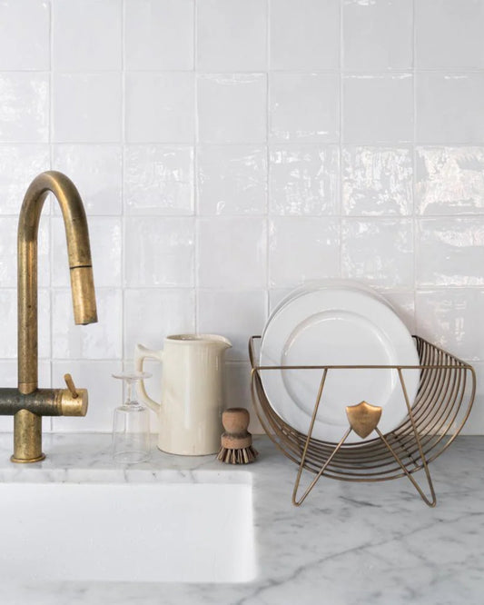 A marble countertop with a corrosion-resistant brass faucet showcases the COMPASS DISH RACK by Society Ink, holding white plates and a beige pitcher. Nearby, a clear glass and a small scrub brush lean against the white tiled backsplash.