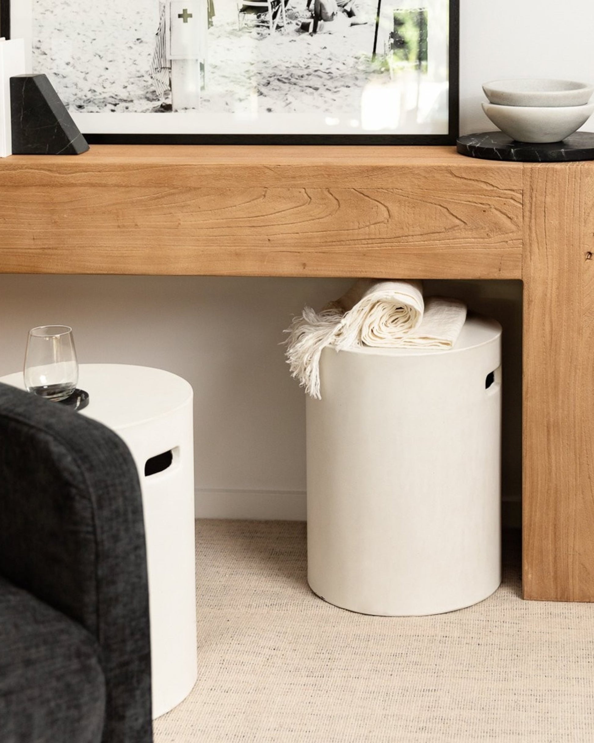 A cozy living room corner features a large wooden console table that emphasizes minimalist design. A rolled-up white blanket rests on a Hawthorne CONCRETE PIPE SIDE TABLE / STOOL, while another identical stool nearby holds a glass and a bowl. Above, an elegant touch is added by a black-and-white framed photo.