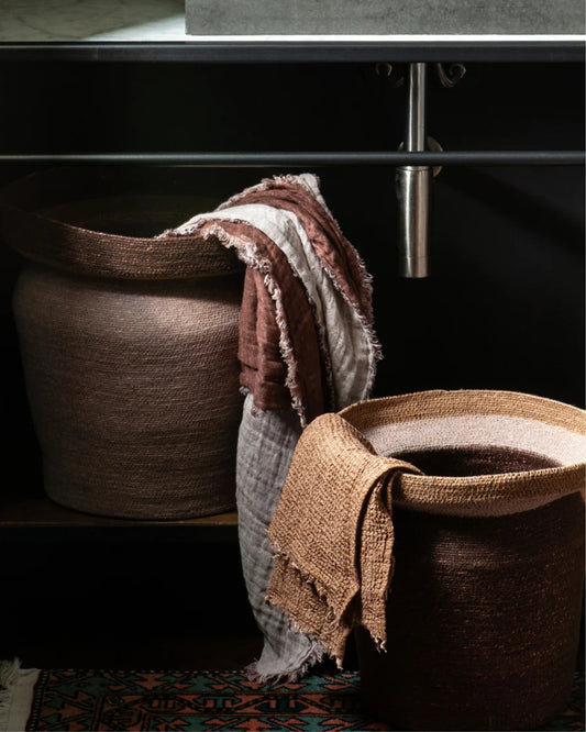 Two medium-sized CONTENERE CACAO baskets by Bianca Lorenne, featuring textured brown and cream cloths draped over them, are placed on a dark shelf beneath a metallic pipe. Below, a partially visible patterned rug enhances the space with its vibrant colors, creating an organic storage scene.