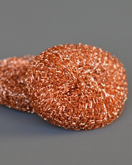Two Redecker Copper Scourers rest on a smooth gray surface. The scourers feature a shiny, woven texture made of copper fibers and are perfect for cleaning tasks. Additionally, they are recyclable, offering both practicality and environmental friendliness.