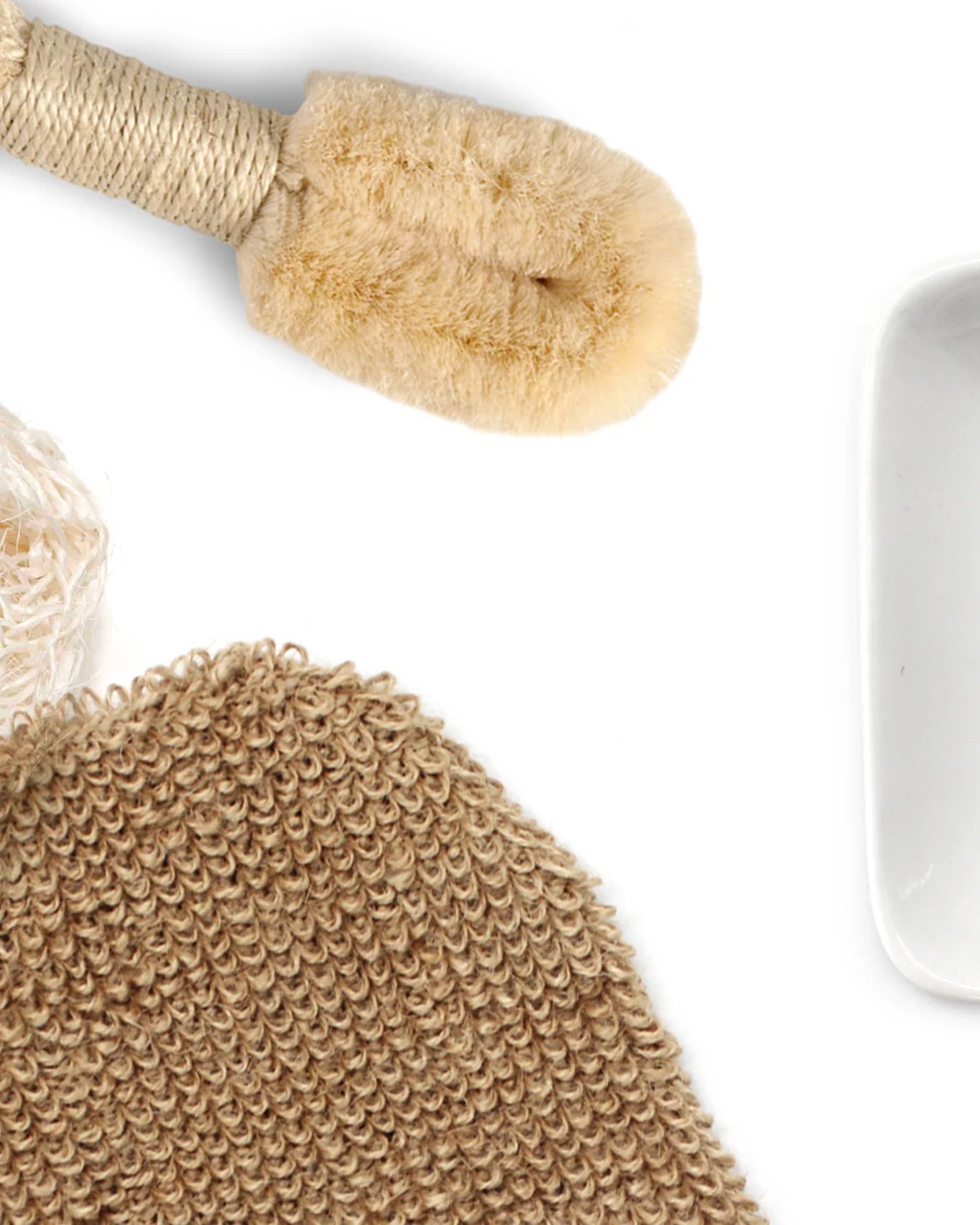 A CORSICA BATH MITT from Saison, featuring natural bristles and a handle woven from sisal fibre, is artfully arranged alongside a beige knitted exfoliating scrubber and a white ceramic dish on a white background.