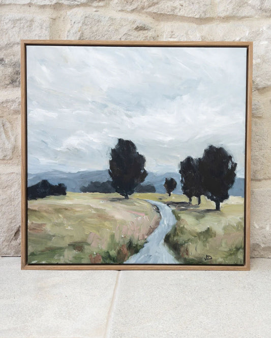 The "COUNTRY MEADOWS - ART BY JEANNIE" framed painting by the intuitive artist Jeannie Dolan exquisitely captures a serene landscape featuring a winding path through grassy fields. The scene includes trees on either side under a cloudy sky, with distant hills visible in the background, beautifully reflecting the natural world against a light stone wall backdrop.