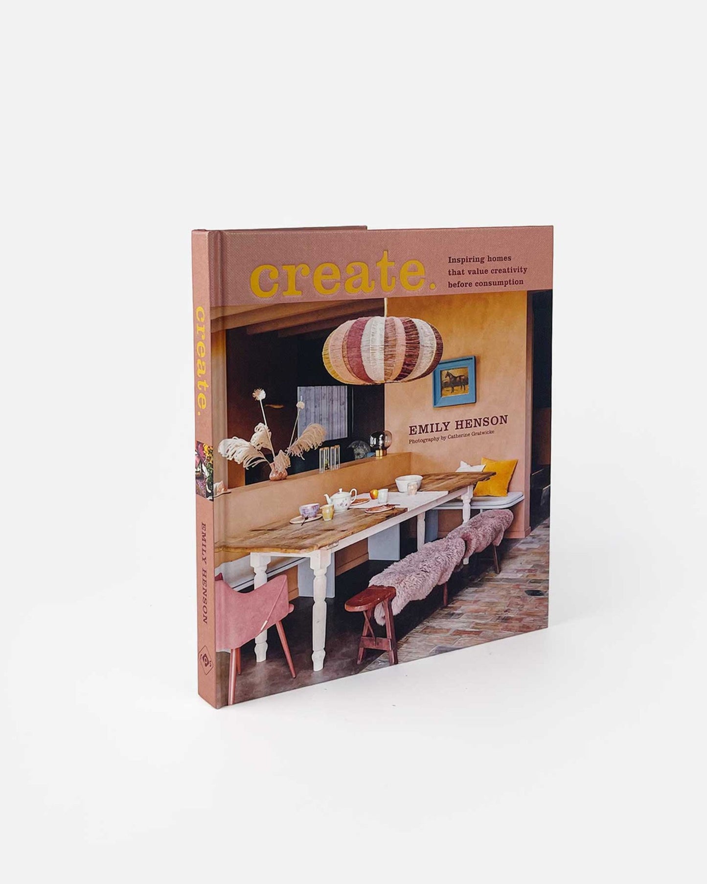 The image depicts a book titled "CREATE: INSPIRING HOMES" by Bookreps. The cover showcases a chic dining room with a long table adorned with decor items and pink cushions. The book emphasizes embracing creativity and individuality in home design through personal style and repurposed pieces.