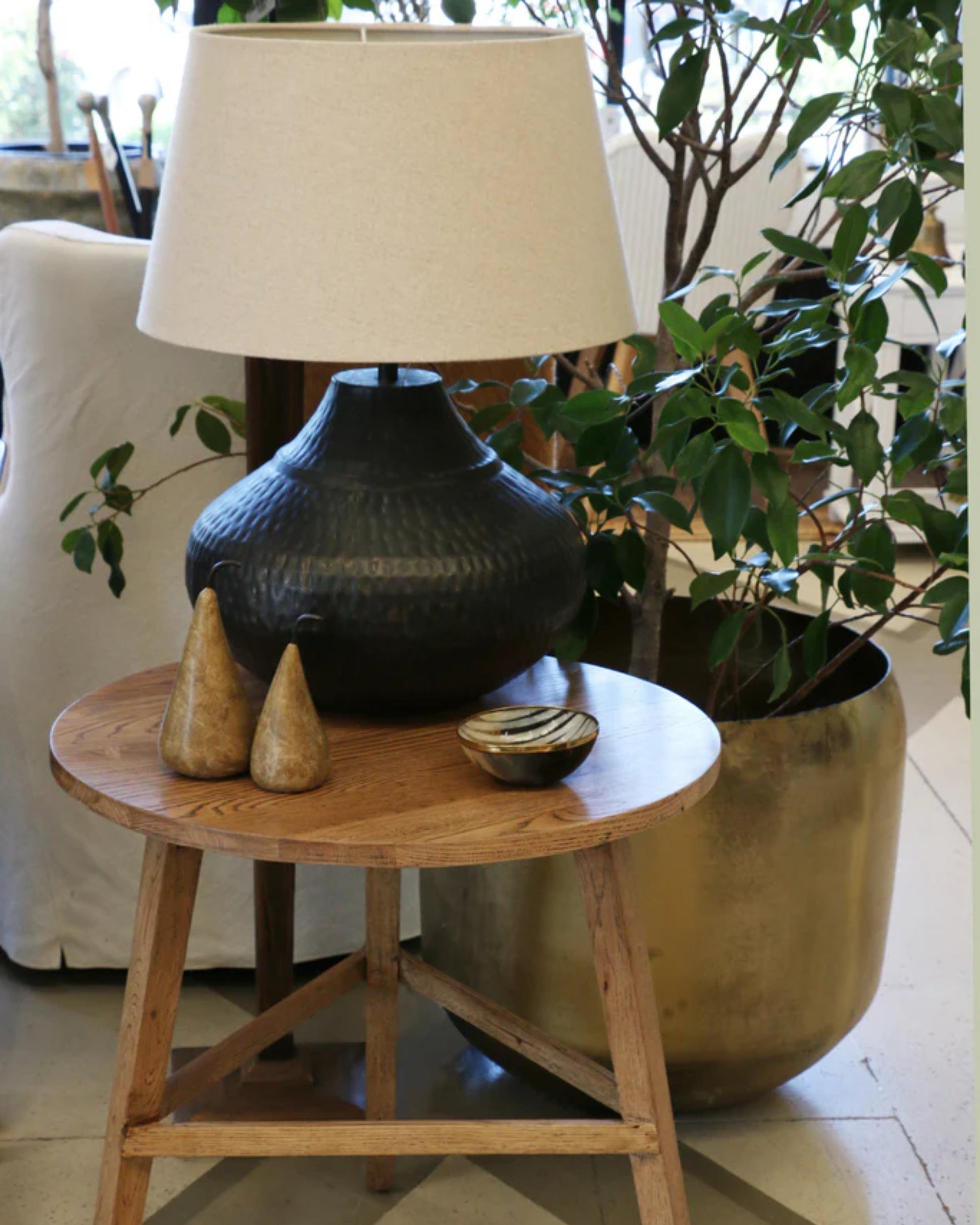 A cozy living room scene features a CRICKET TABLE by CC Interiors, showcasing a textured black lamp with a beige shade, two decorative pear-shaped objects, and a small dish. Nearby, a large potted plant adds greenery to the space.