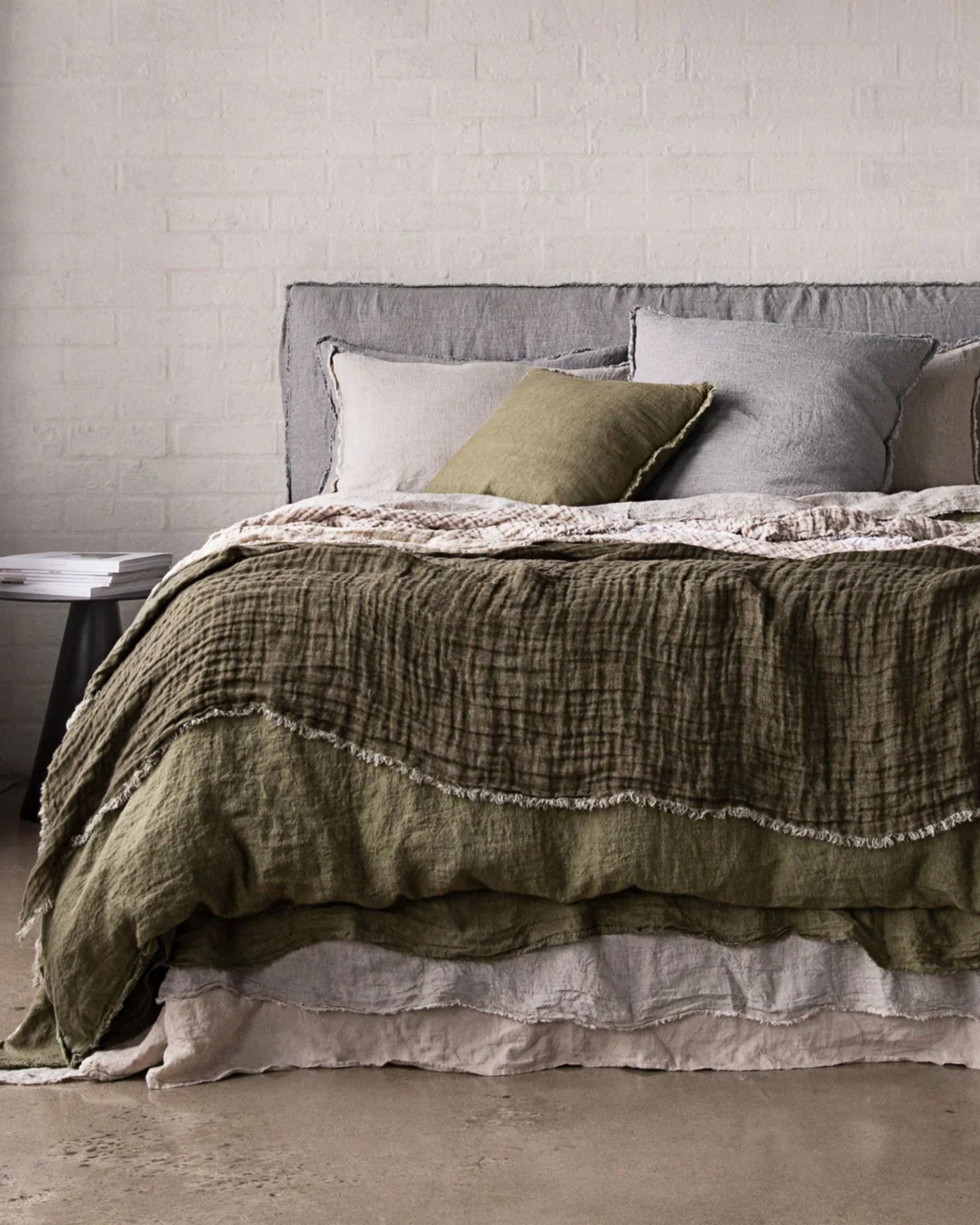 A cozy bed with a luxurious accent from an olive green, pure linen CRUSH DOUBLE LINEN THROW BLANKET in Armee/Sable by Hale Mercantile Co and several pillows against a light grayish-brick wall. A small round side table with books is positioned next to the bed on a wooden floor, enhancing the room's calm, minimalist decor.