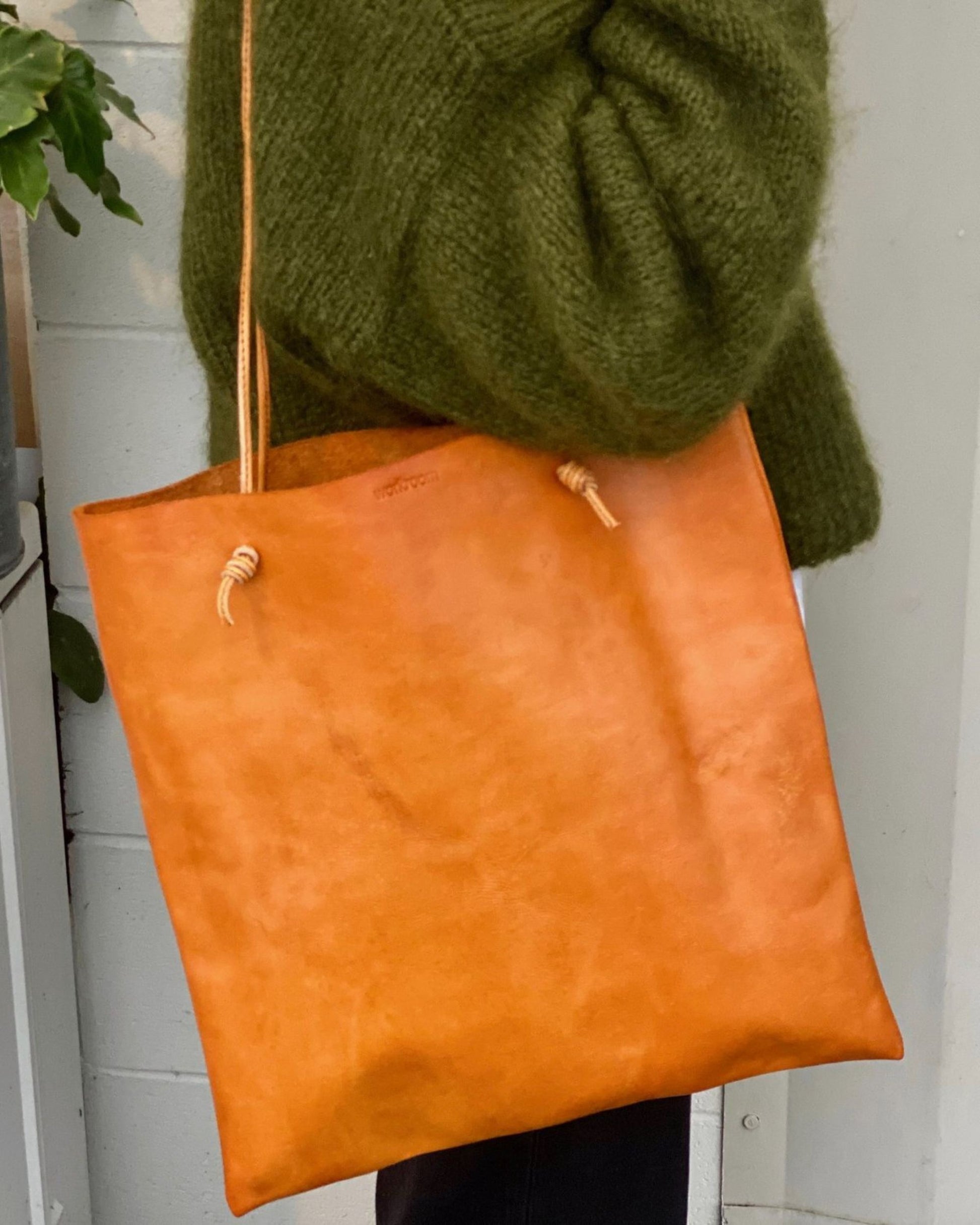 A person holds The Workroom's CUIR KNOT TOTE BAG, showcasing its simple design, while wearing a green sweater. The light-colored background includes part of a plant.