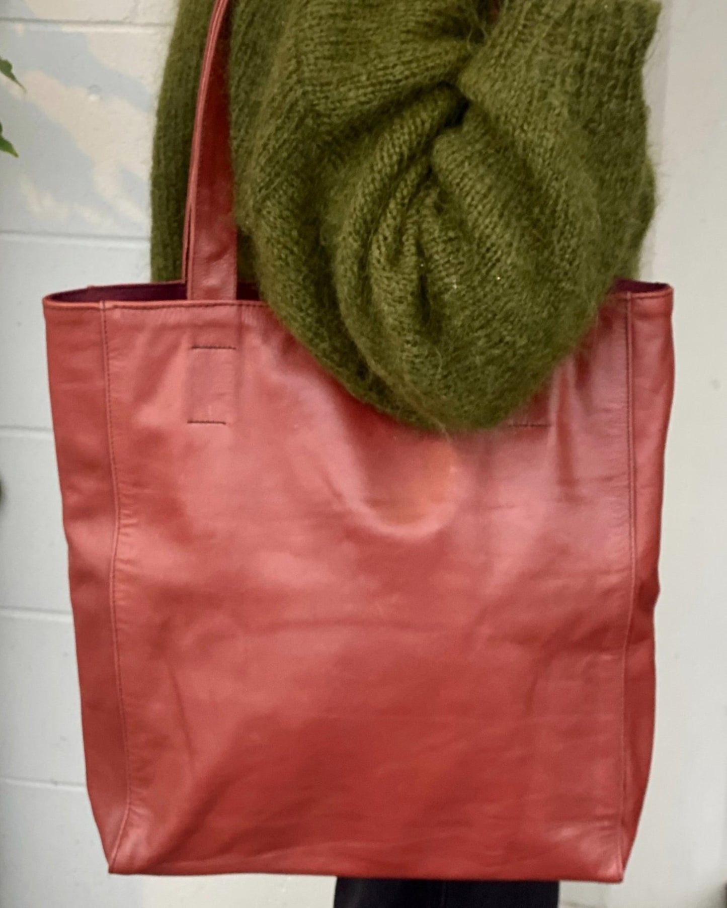 A person wearing a thick green sweater holds the CUIR LINED TOTE BAG, a stylish and soft leather creation by The Workroom. The background is a neutral-colored wall.