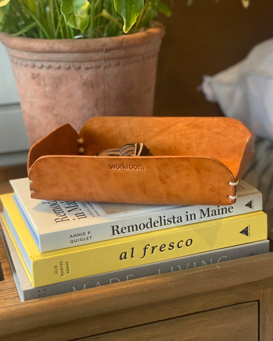 Books are artfully arranged on a wooden surface with The Workroom's CUIR LEATHER TRINKET TRAY elegantly placed on top. Titles such as Remodelista in Maine and al fresco, among others, complement this luxurious storage piece. In the background, a plant in a terracotta pot peeks into view.