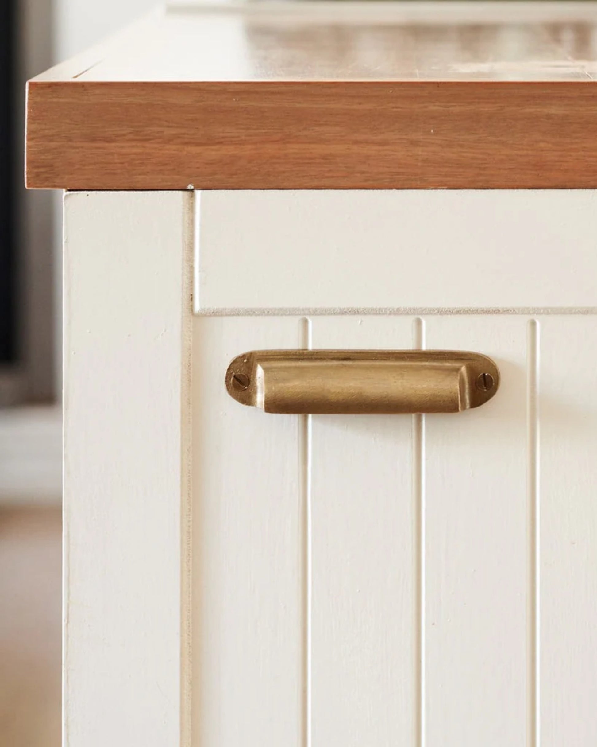 Close-up of a white cabinet with vertical panel detailing, featuring Society Ink's CURIO DRAWER PULL in brass and a light wood countertop, designed in Australia.