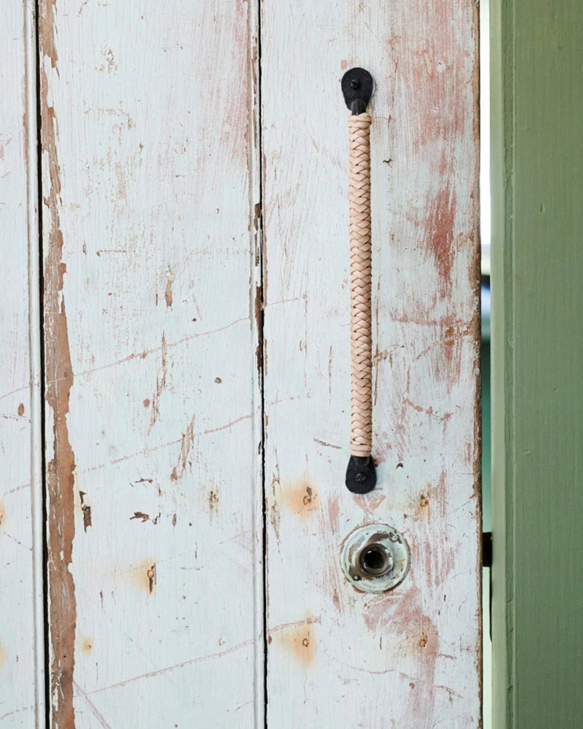 Close-up of a rustic wooden door with chipped white paint, featuring Society Ink's CUTTER PULL—a braided rope handle attached with metal fixtures. An empty round doorknob hole is visible, while aged leather accents resemble drawer pulls on the right side's green door frame.