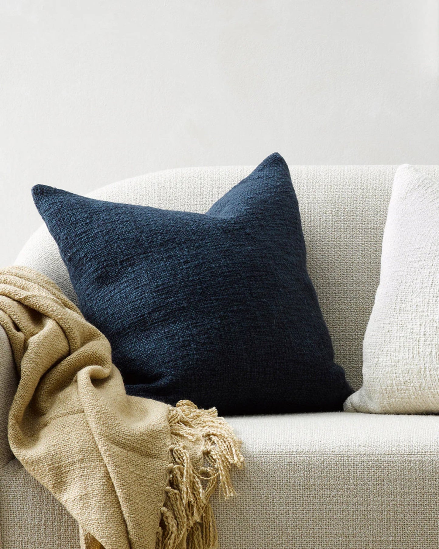 A cozy white couch featuring the CYPRIAN CUSHION by Baya in a dark blue textured design, along with a handwoven beige fringed throw blanket elegantly draped over the armrest. The neutral off-white wall in the background enhances the rustic texture of the setting.