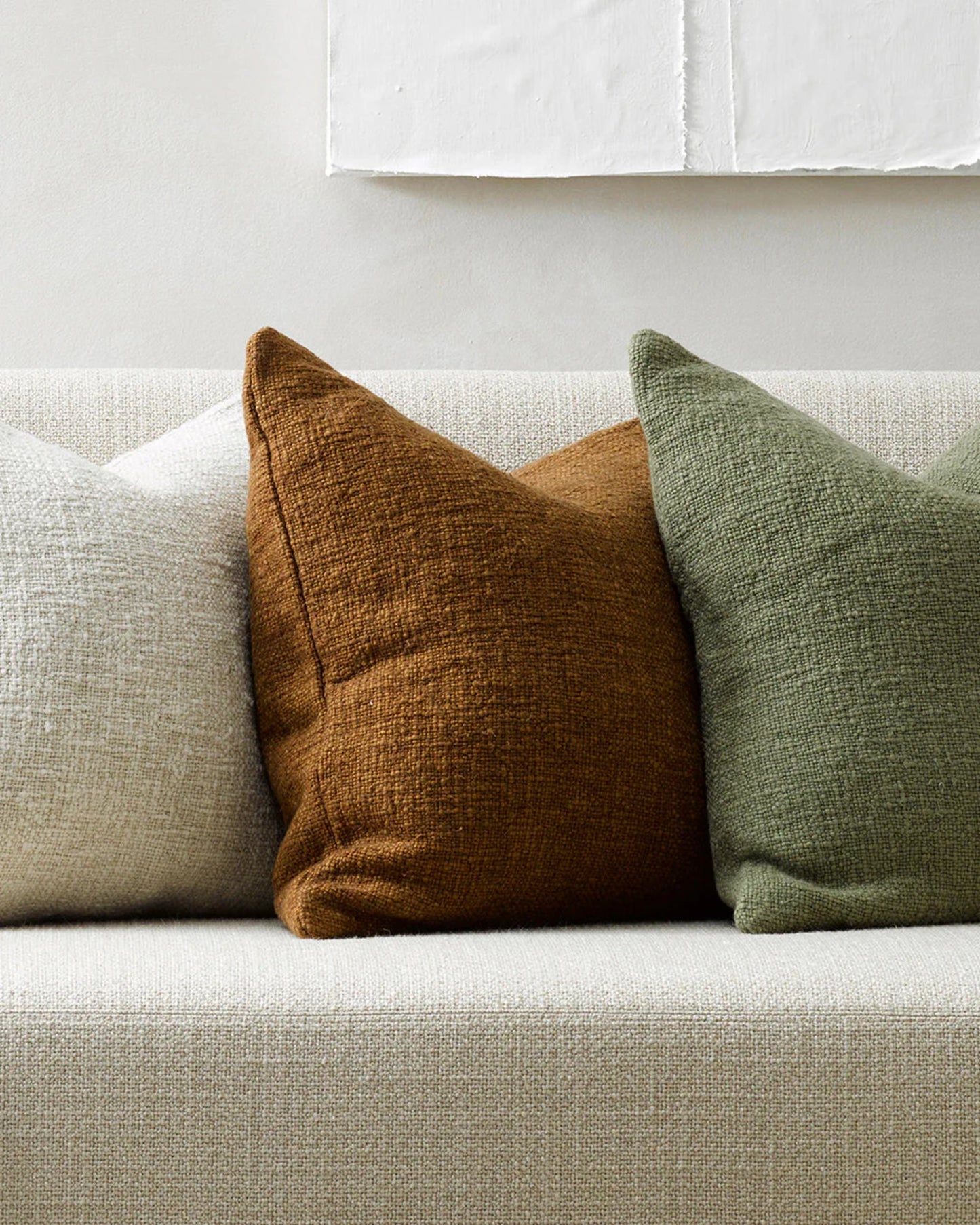 A white couch is styled with a line of three CYPRIAN CUSHIONS by Baya, showcasing a textured design in cream, brown, and forest green. The wall behind is light-colored, featuring handwoven white fabric art above. This minimalistic scene exudes coziness and rustic charm.