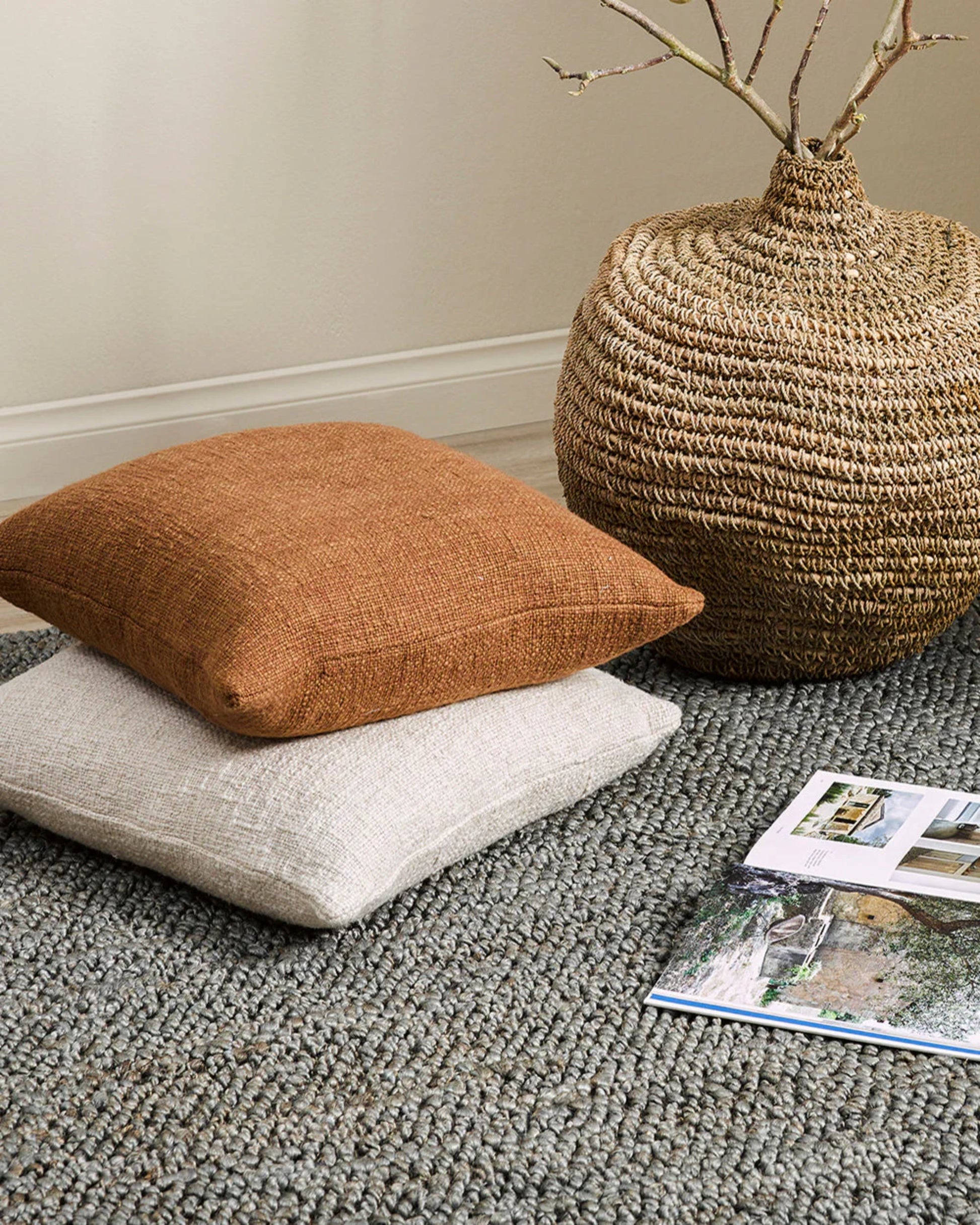 A pair of CYPRIAN CUSHIONS by Baya, one in a brown texture and the other in beige, are neatly arranged on a gray woven rug. Close by, a wicker basket filled with twigs and an open magazine enhance the cozy, rustic ambiance of the interior setting.