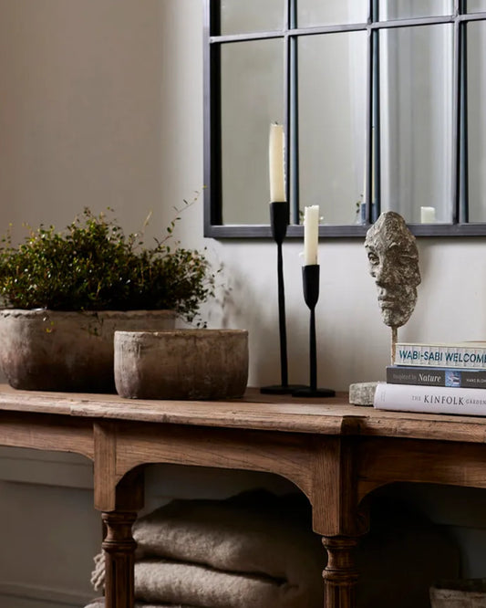 A rustic wooden table with a potted plant and a stone bust sits beneath a grid-framed mirror. The decorative setup is enhanced by two DAX CANDLEHOLDERS in matte black by French Country and a stack of books, creating a cozy and artistic ambiance.