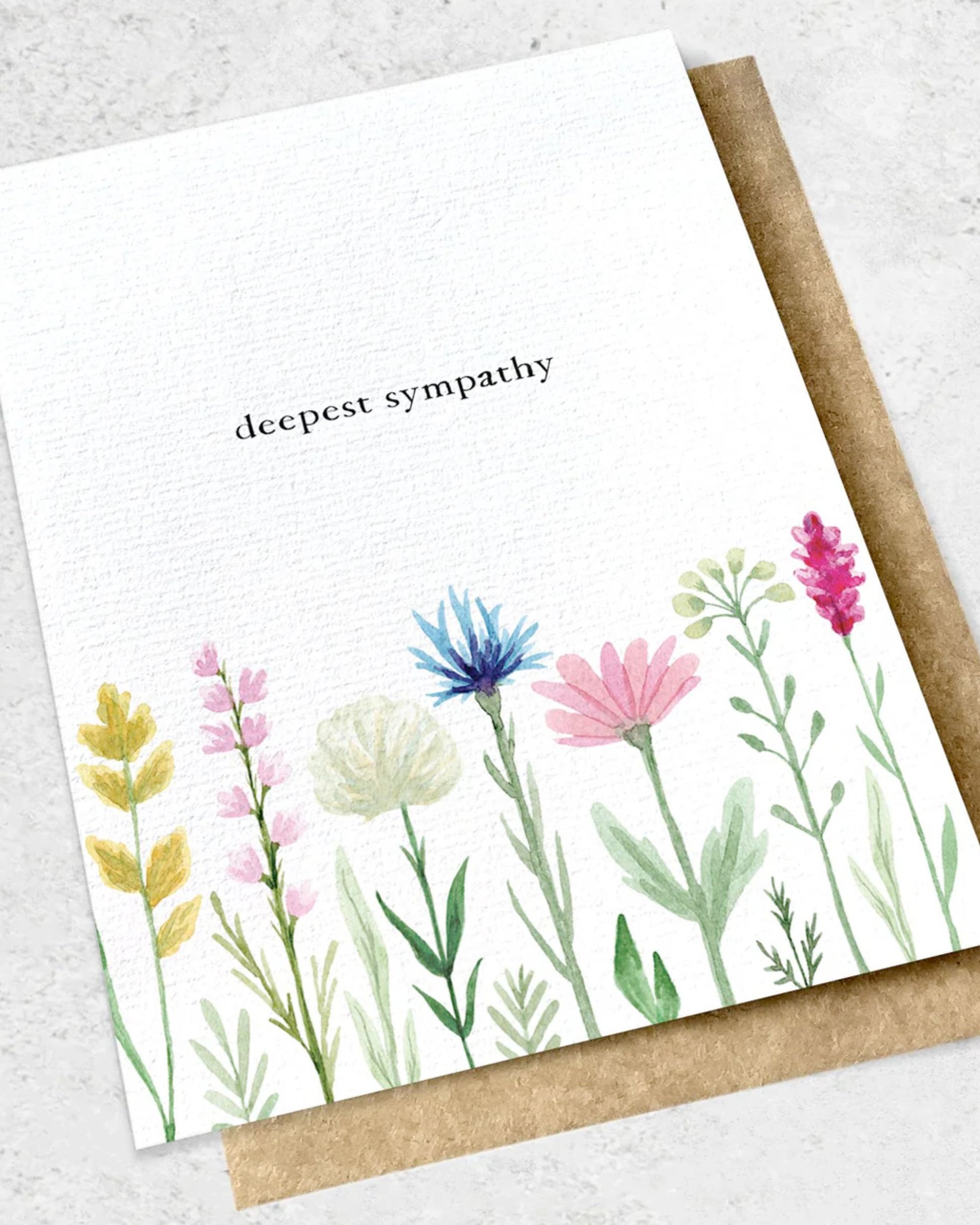 Product Description: The "DEEPEST SYMPATHY FLOWERS - GREETING CARD" by Ink Bomb features the words "deepest sympathy" surrounded by delicate watercolor flowers in pink, blue, yellow, and green along the bottom. This premium textured card comes with a kraft envelope and is elegantly presented against a textured backdrop.