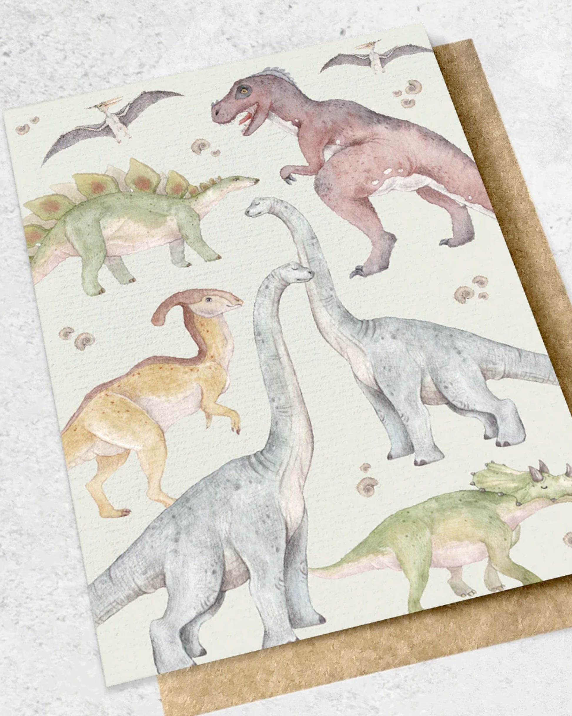 Introducing the DINOSAUR - GREETING CARD by Ink Bomb, showcasing a delightful illustration of dinosaurs such as Tyrannosaurus rex, Stegosaurus, Brachiosaurus, and Triceratops with pterodactyls gracefully flying above. This premium textured card features a light background and is perfectly complemented by its kraft envelope.