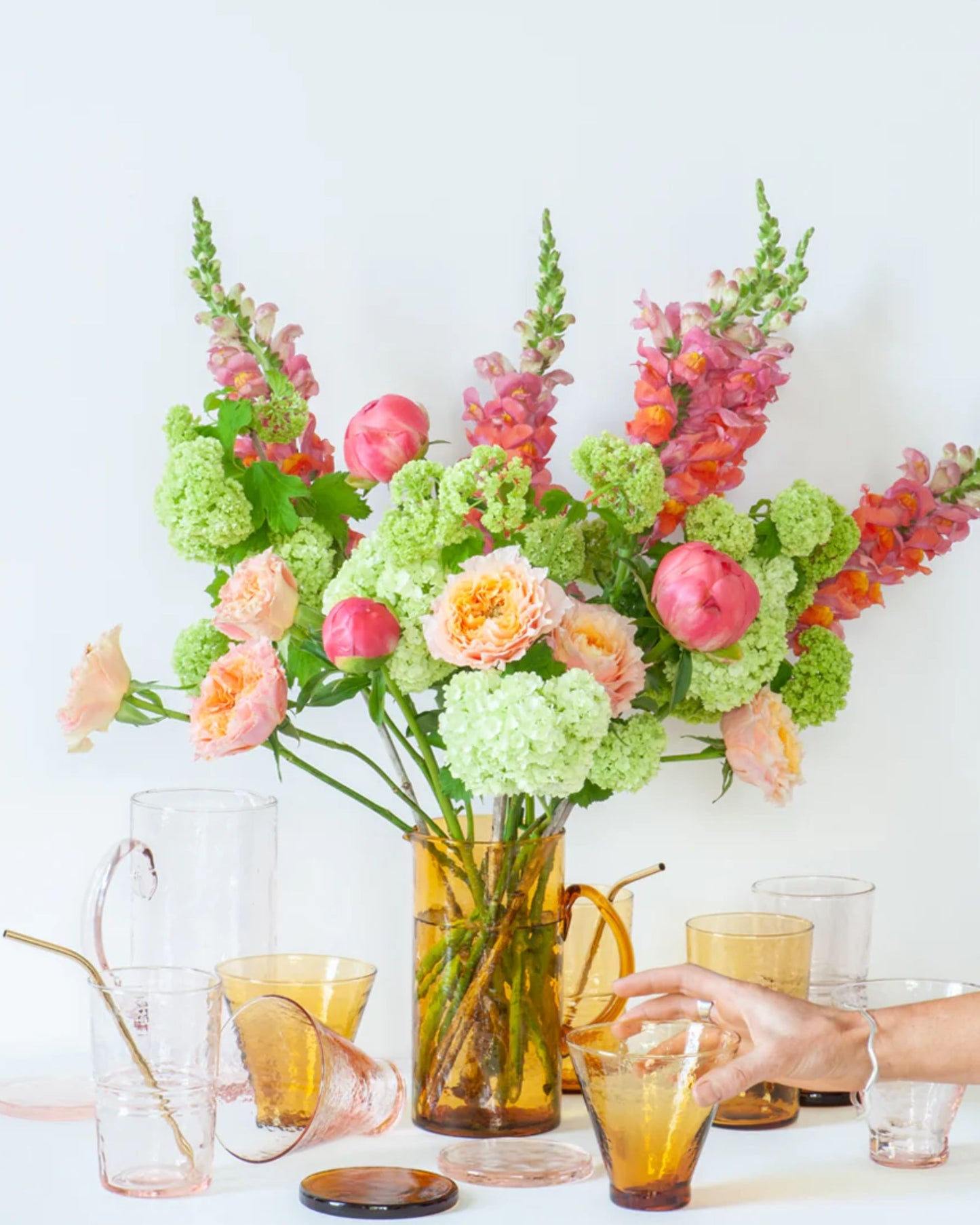 An exquisite floral arrangement bursting with pink, peach, and green flowers graces an amber vase. Surrounding this display are various pieces of Bianca Lorenne's handmade glassware. A hand reaches for one of the DRINKING GLASSES - AMBER from the diverse selection on the table, perfectly highlighting the aesthetic charm of recycled glass in this beautiful amber colorway.