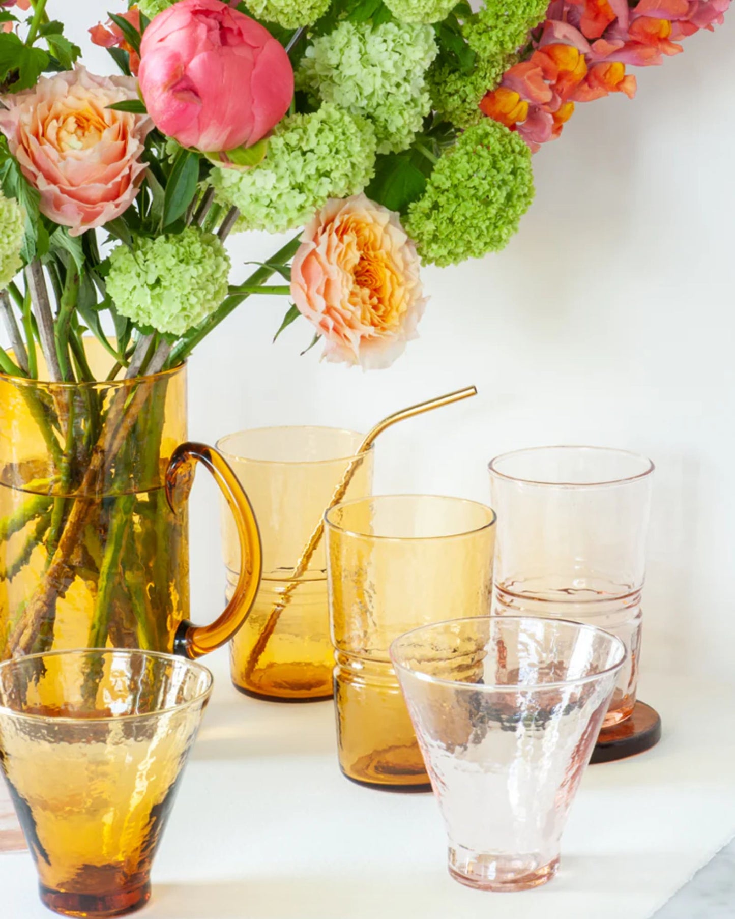 A vibrant bouquet with pink and orange flowers is beautifully displayed in an amber glass pitcher, highlighting the signature colorway of Bianca Lorenne's DRINKING GLASSES - AMBER. Beside it, a selection of handmade recycled glassware, including cups and a tumbler with a straw, is tastefully arranged on a pristine white surface.