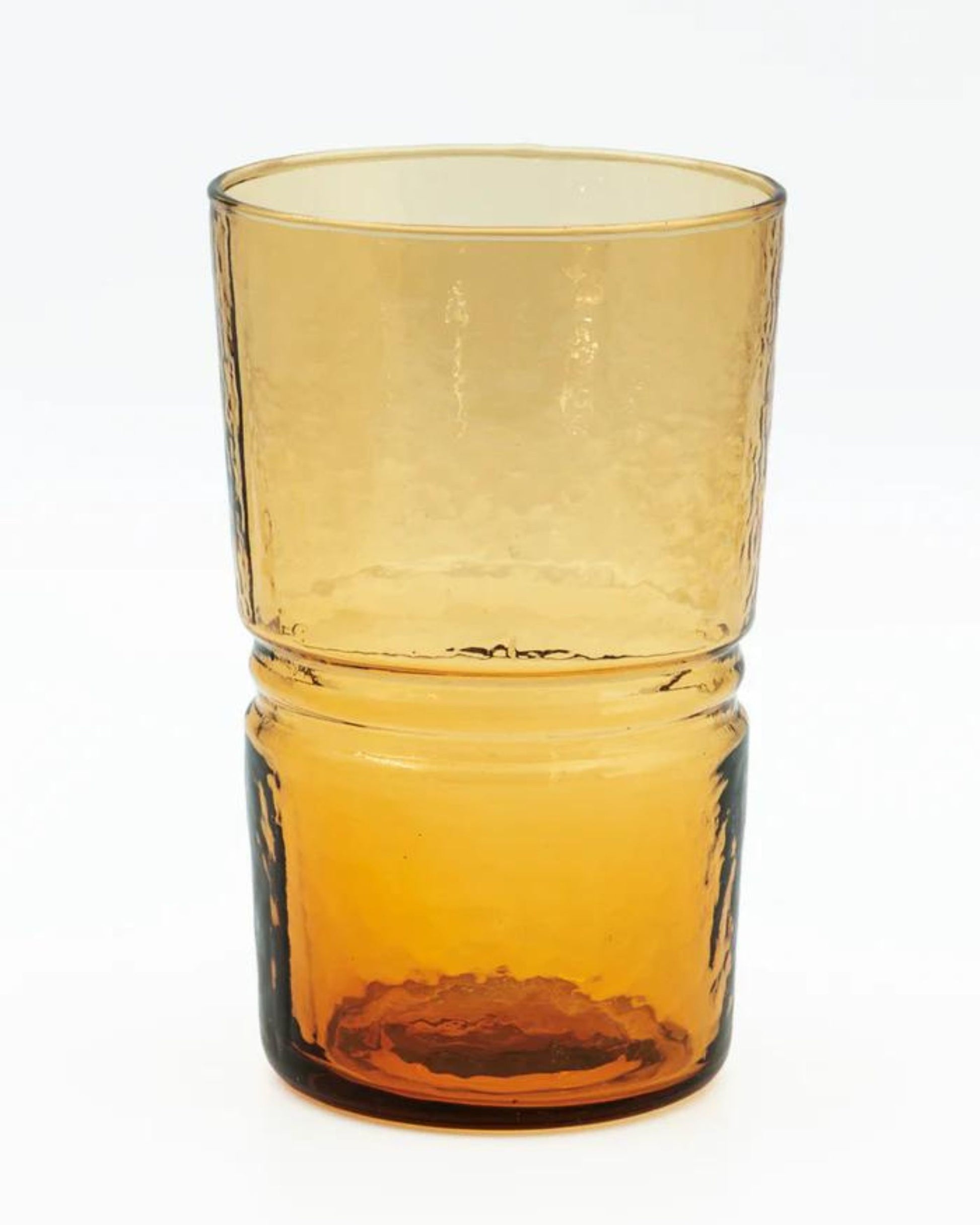 The DRINKING GLASSES - AMBER by Bianca Lorenne features a translucent amber glass with a textured surface and a slightly tapered design. This handmade glassware boasts a thicker base and smooth, rounded edges, skillfully crafted from recycled glass. The product is displayed against a plain white background.