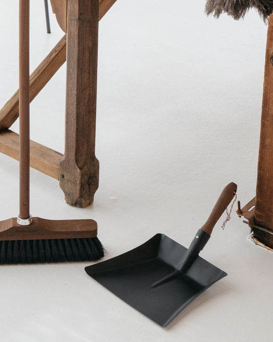 A wooden broom with a French beechwood handle rests next to an Andree Jardin DUSTPAN made of black sheet metal on a white floor, complemented by elegant wooden furniture.