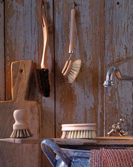 Saison's ECO BEECH WOOD DISH BRUSH, featuring tampico fibre bristles, hangs on a rustic wooden wall in the kitchen. Above the sink, a metal faucet is accompanied by a textured cloth draped over the edge. Nearby, a round dish brush and a sustainable short-handled brush are neatly placed on the shelf.
