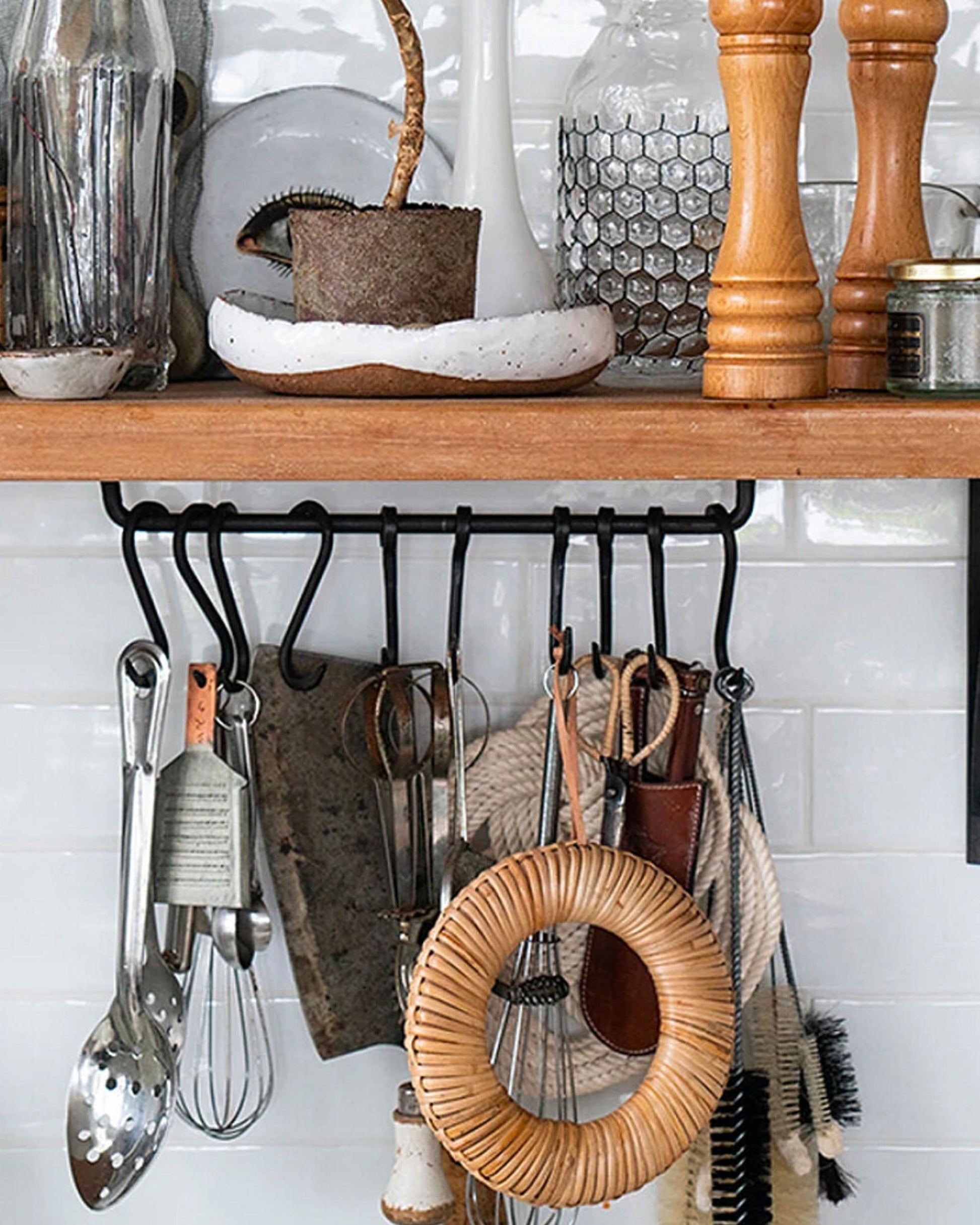 A kitchen shelf captures the essence of wabi-sabi with its delightful assortment of utensils and decor. Hanging items such as whisks and slotted spoons are complemented by the Society Ink EDO CANE TRIVET, while a pot with a plant, glass bottles, and wooden salt and pepper mills are set against a white tiled backsplash.