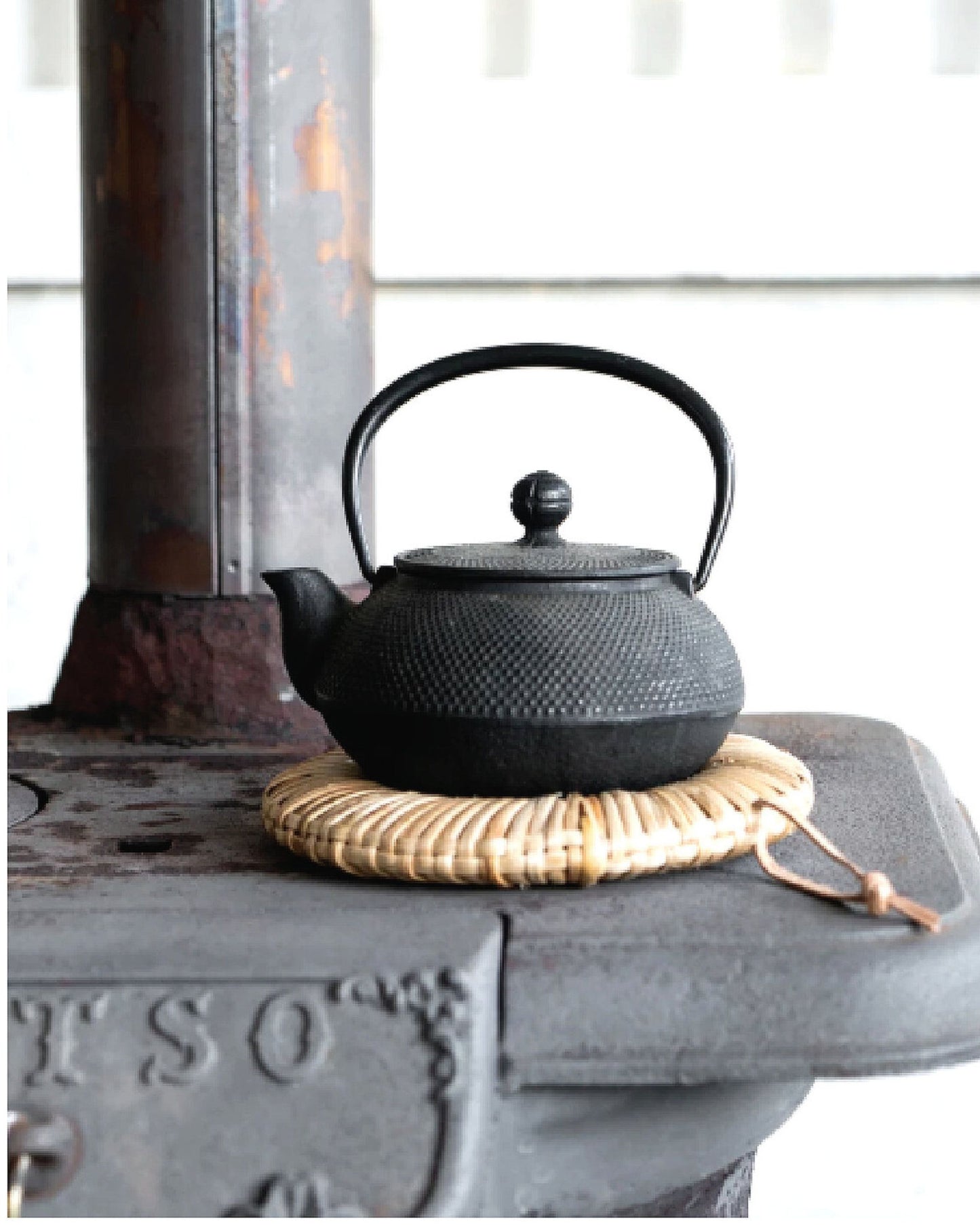 A black cast iron teapot with a textured surface rests on a Society Ink EDO CANE TRIVET atop an antique stove. The stove's rustic, weathered appearance enhances the wabi-sabi aesthetic of the setting.