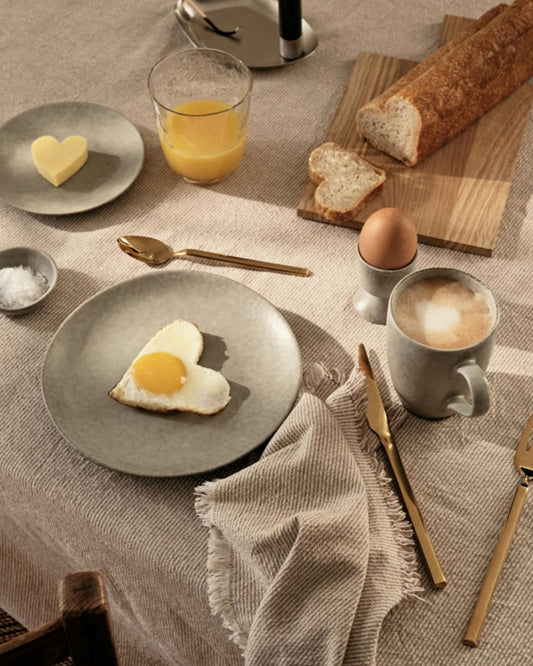 A cozy breakfast setup on a beige tablecloth showcases a heart-shaped fried egg on a plate, a slice of butter, and an elegant Broste EGG CUP holding a soft-boiled egg. Accompanying the scene are glasses of orange juice and coffee, along with a fresh baguette. Gold cutlery and a fringed napkin complete the arrangement on this charming breakfast table.