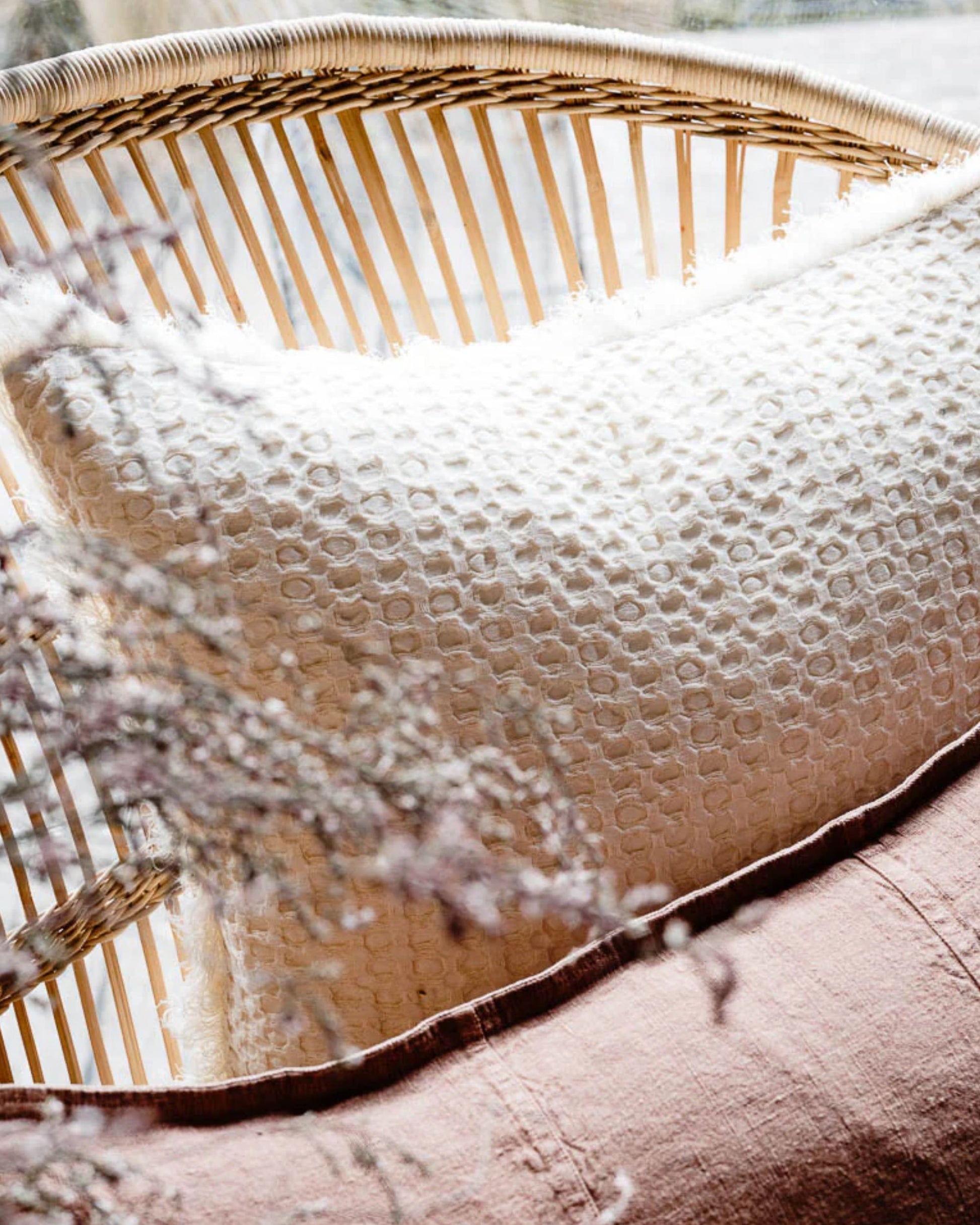 A cozy indoor scene features the ELEMENT CUSHION - IVORY 50X50 by Eadie alongside another textured pillow in mauve, both crafted from a luxurious linen-cotton blend, resting on a wicker chair. Soft sunlight filters through the window, casting gentle shadows and enhancing the boho effect of this warm, inviting setting.