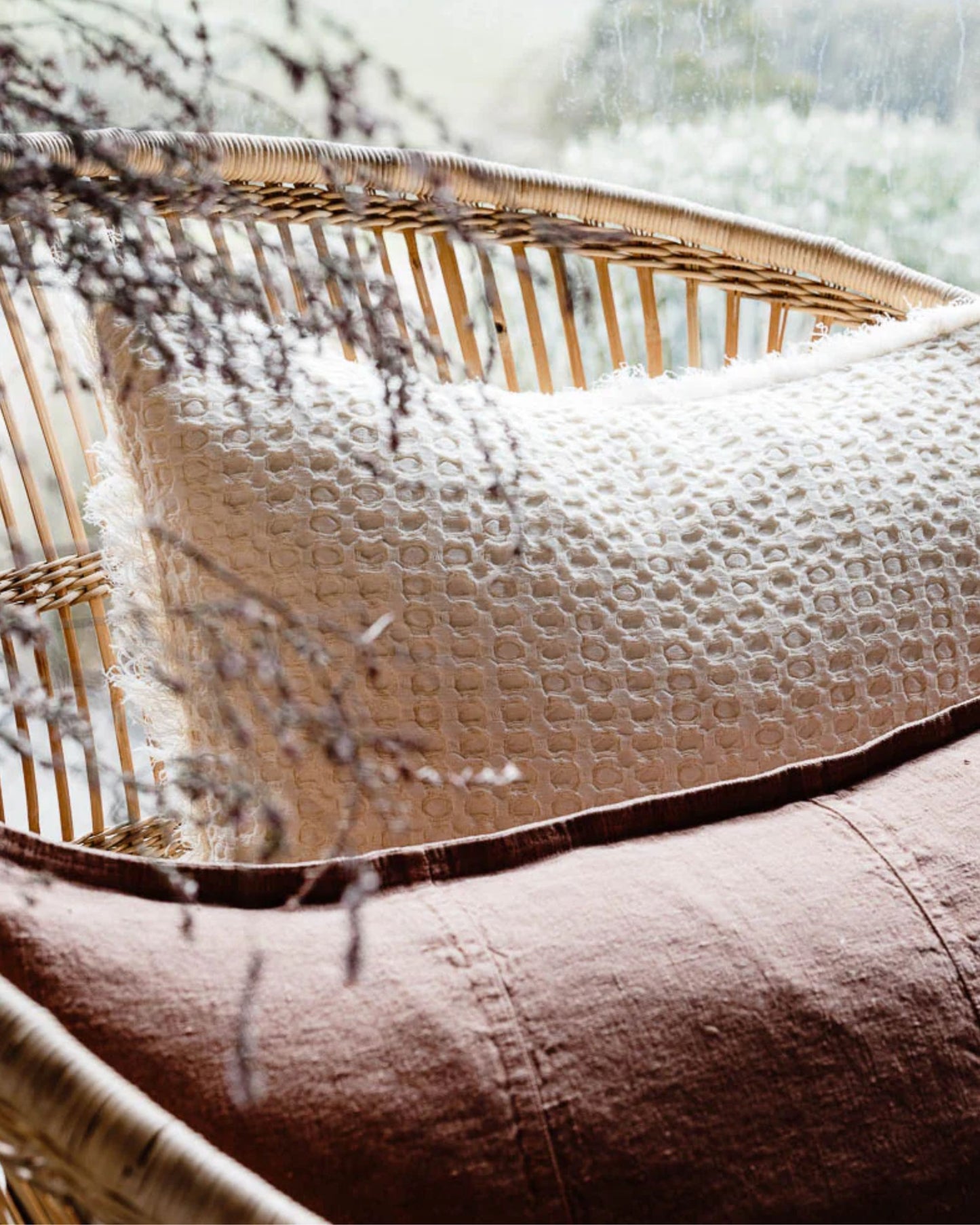 A wicker chair featuring the ELEMENT CUSHION - IVORY 40x60 from Eadie, showcasing a cream-colored cushion with an ivory circular pattern and a solid brown textured cushion in a boho style. Dried branches partially frame the scene, while soft natural light streams through a window in the background.