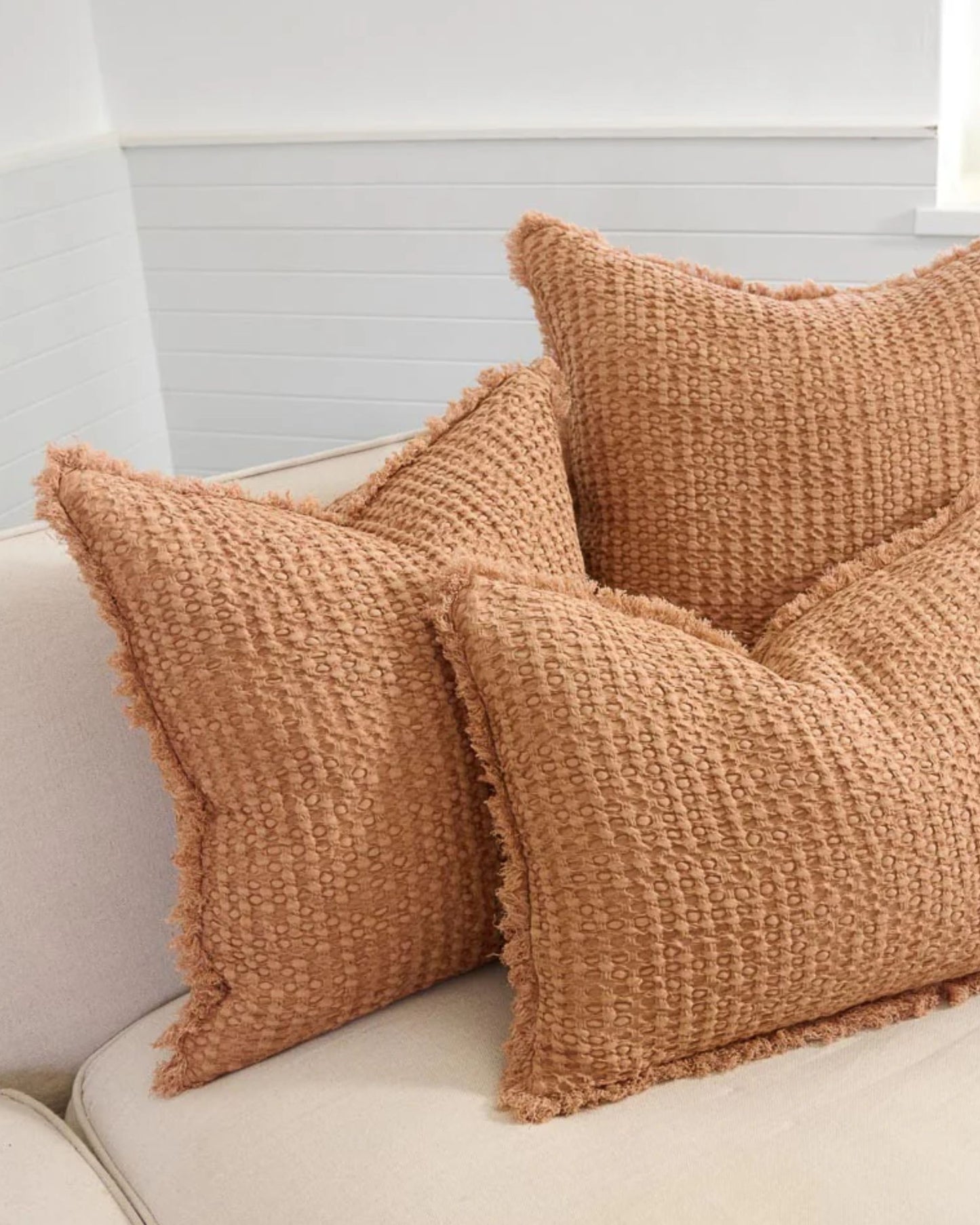 Three Element Cushions in Ochre, each measuring 40x60 and crafted by Eadie with a linen cotton blend, are arranged on a light beige sofa. These cushions feature a woven appearance with fringed edges and are set in a bright, minimalistic room with white shiplap walls.