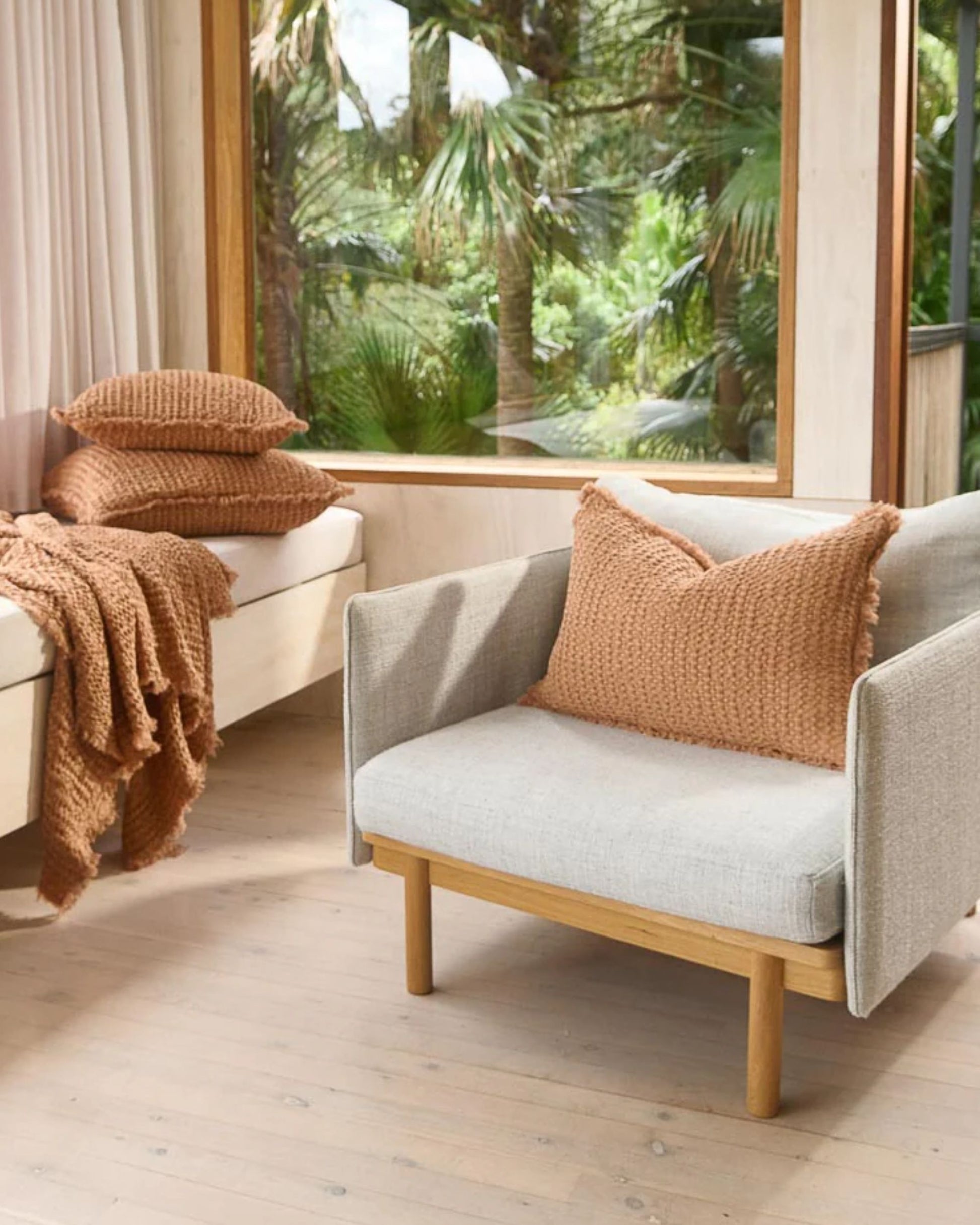 A cozy room with a large window showcasing lush greenery outside. Inside, the boho effect is brought to life with a gray armchair decorated with Eadie’s Element Cushion - Orchre 40x60, made from a textured linen cotton blend in orange. A wooden bench complements the setting, featuring matching throw pillows and a blanket on the light wooden floor.