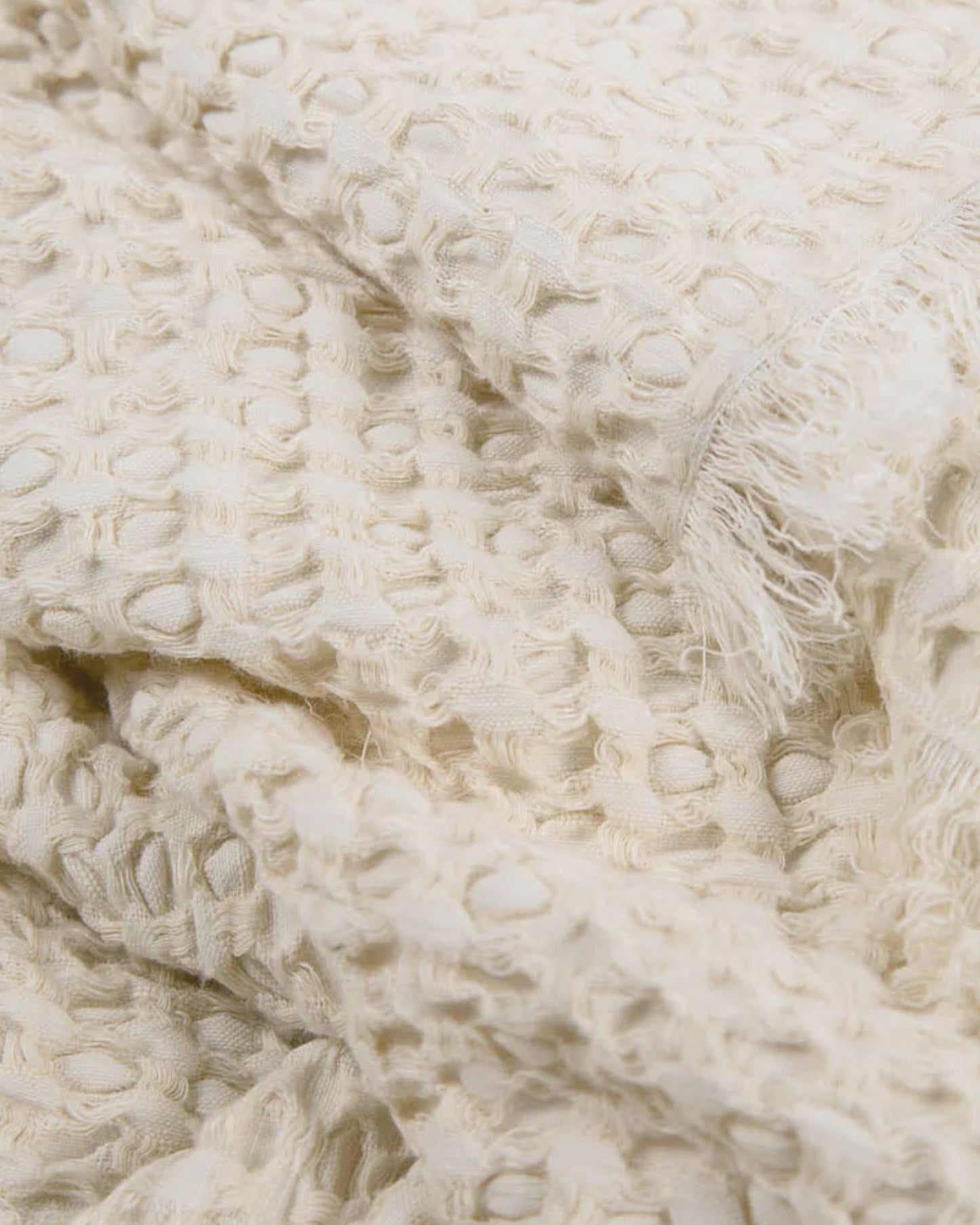 A detailed view of Eadie's ELEMENT THROW - IVORY reveals its textured fabric with a waffle pattern and fringed edges, highlighting its soft and cozy feel accented by a boho effect.