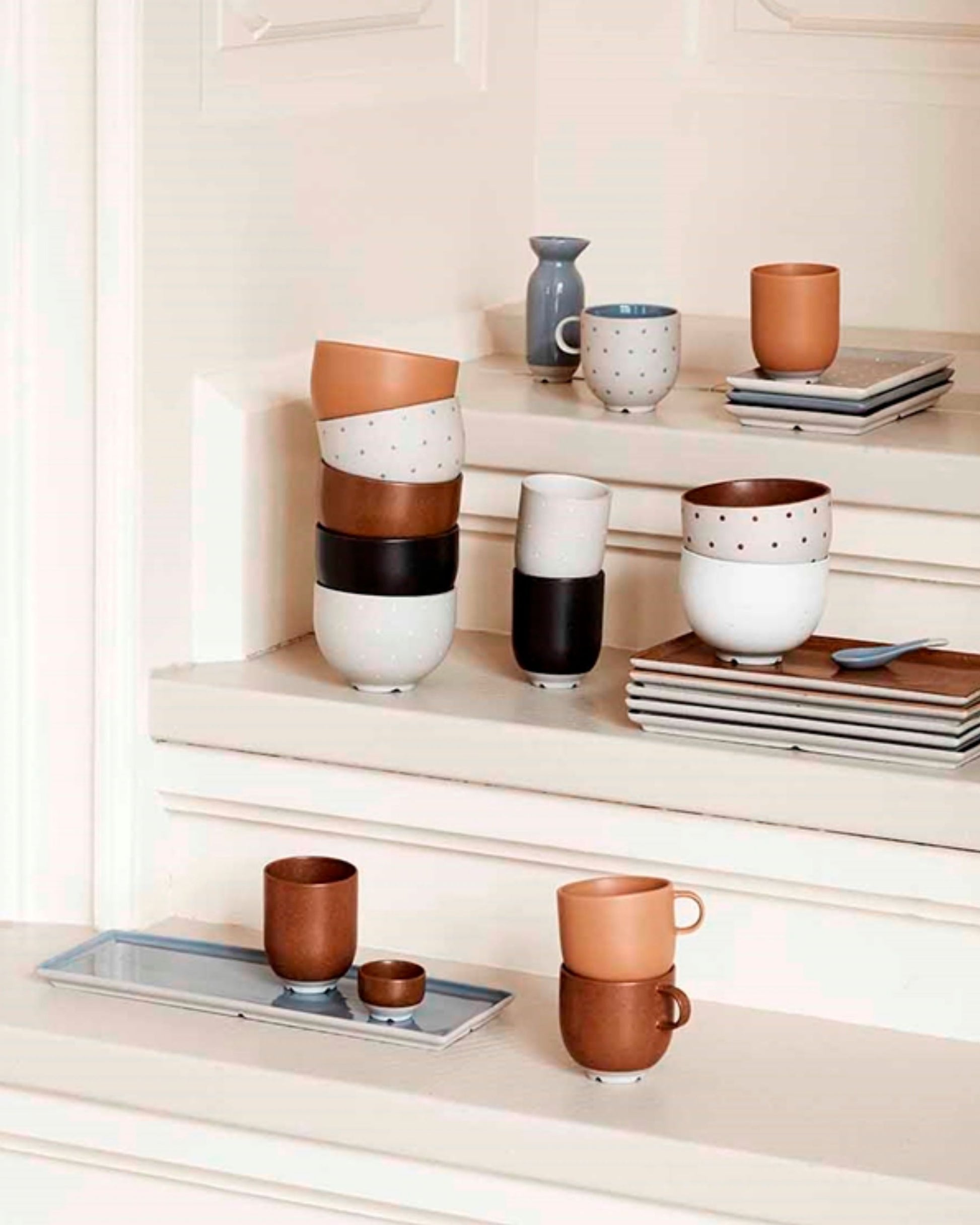 Assorted ceramic items, including the ELI MINI BOWL by Broste, showcase a reactive glaze in shades of brown and white with speckled patterns. They are neatly stacked on a white stepped shelf alongside a small blue vase, books, and a tray, creating an aesthetically pleasing arrangement from the Eli collection.