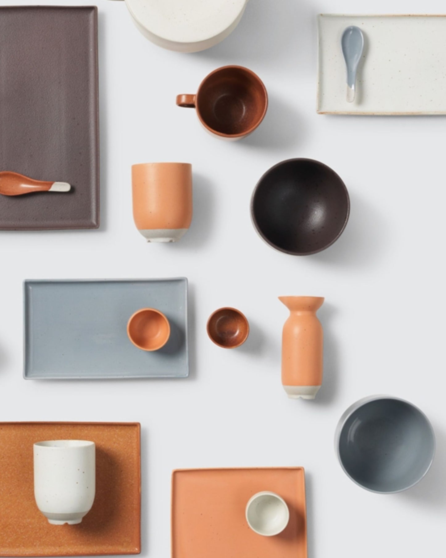 A flat lay showcasing a collection of ceramic tableware, featuring the ELI BOWL in Caramel Brown by Broste, alongside plates, cups, and spoons adorned with a striking reactive glaze. The items are presented in shades of brown, orange, black, white, and gray reminiscent of Japanese tableware and are arranged on a light background.