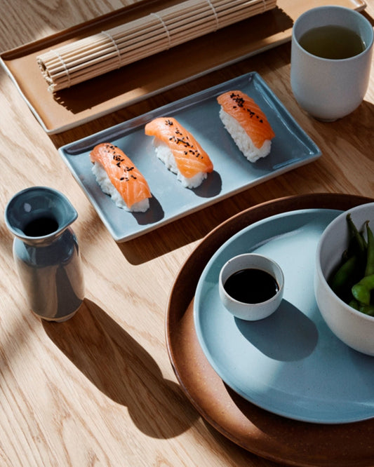 A wooden table is beautifully set with Japanese tableware: three salmon nigiri placed on a rectangular blue plate showcasing a stunning reactive glaze from the Broste Eli collection. Accompanying the plate are a small soy sauce dish, cup, ELI MINI BOWL, soy sauce bottle, and bamboo mat, all illuminated by natural light.