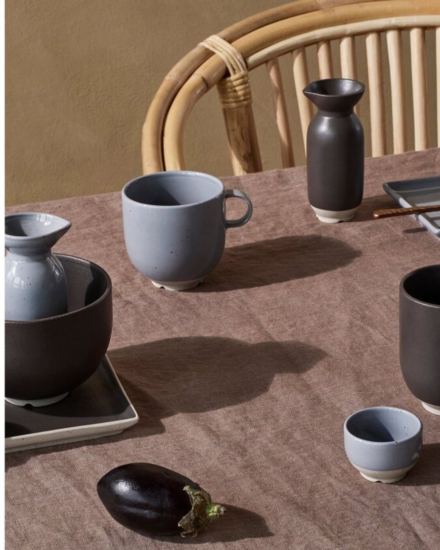 A rustic table arrangement featuring a beige tablecloth highlights the Broste Eli Collection's ceramic mug with handle, bowls, and vase in earthy tones. An eggplant is situated in the foreground, while a wicker chair partially appears, contributing Japanese-inspired elegance to the warm and natural ambiance.