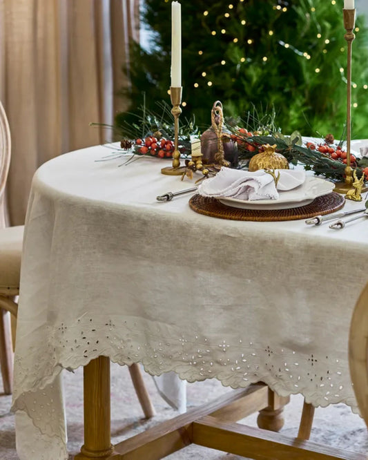 A round table displays elegant holiday settings with a French Country EMBROIDERED TABLE CLOTH WHITE, golden utensils, and a centerpiece adorned with candles, red berries, and greenery. In the background, a Christmas tree sparkles with twinkling lights.