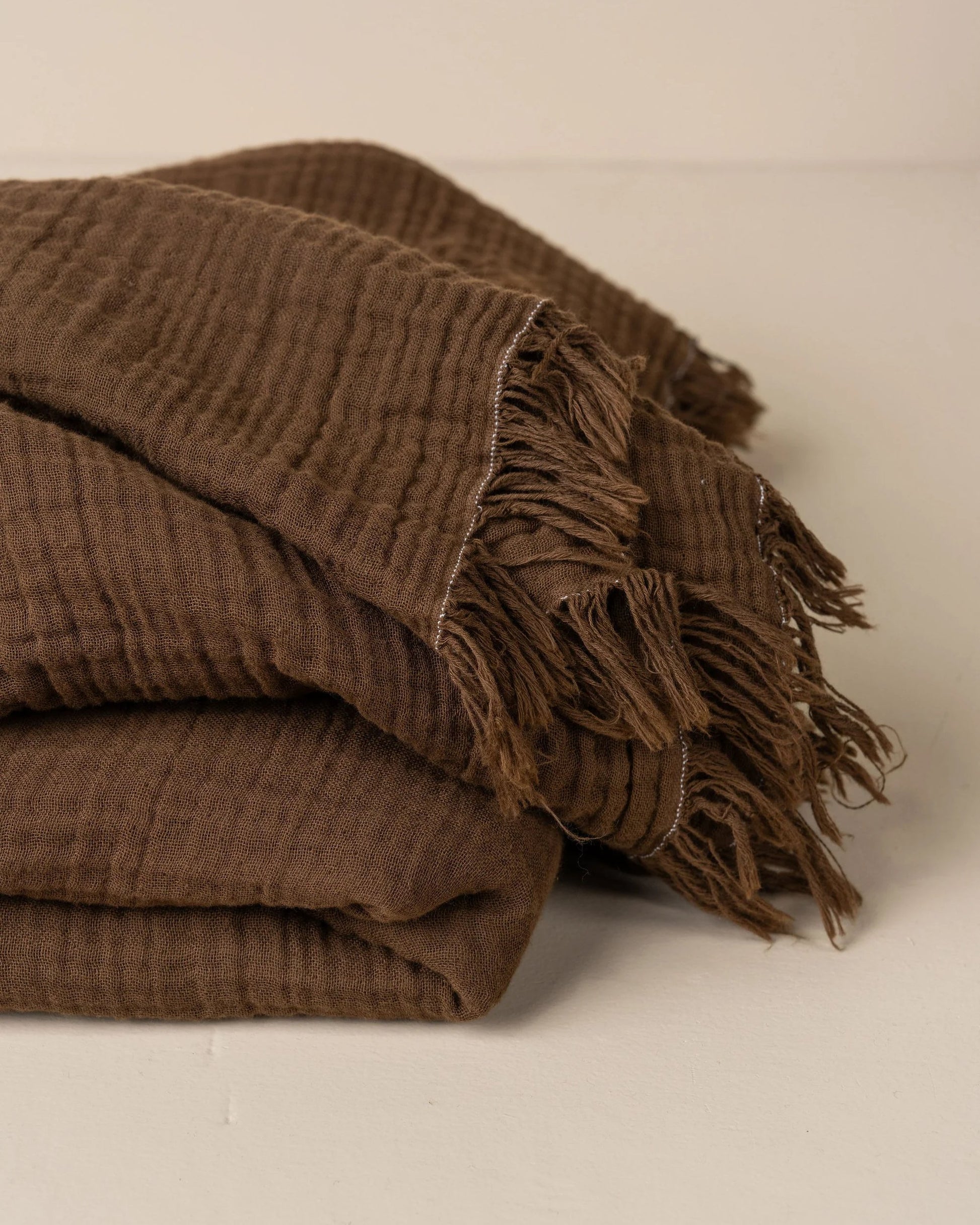 A folded ENES COTTON THROW - CHOCOLATE by Saarde, crafted from crinkled Turkish cotton with fringed edges, rests on a neutral surface. The brown textured throw blanket features a soft and cozy feel, highlighted by its waffle-like pattern, perfect for layering.