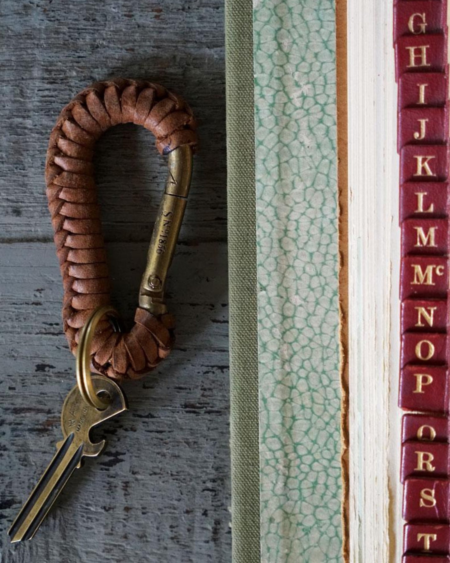 A Society Ink EXPLORER KEYCHAIN with keys is placed next to the spine of a textured book. The book's spine features alphabetically ordered letters on a red background, making it ideal for any adventurer. All of this sits atop a surface that exudes a rustic, worn charm.