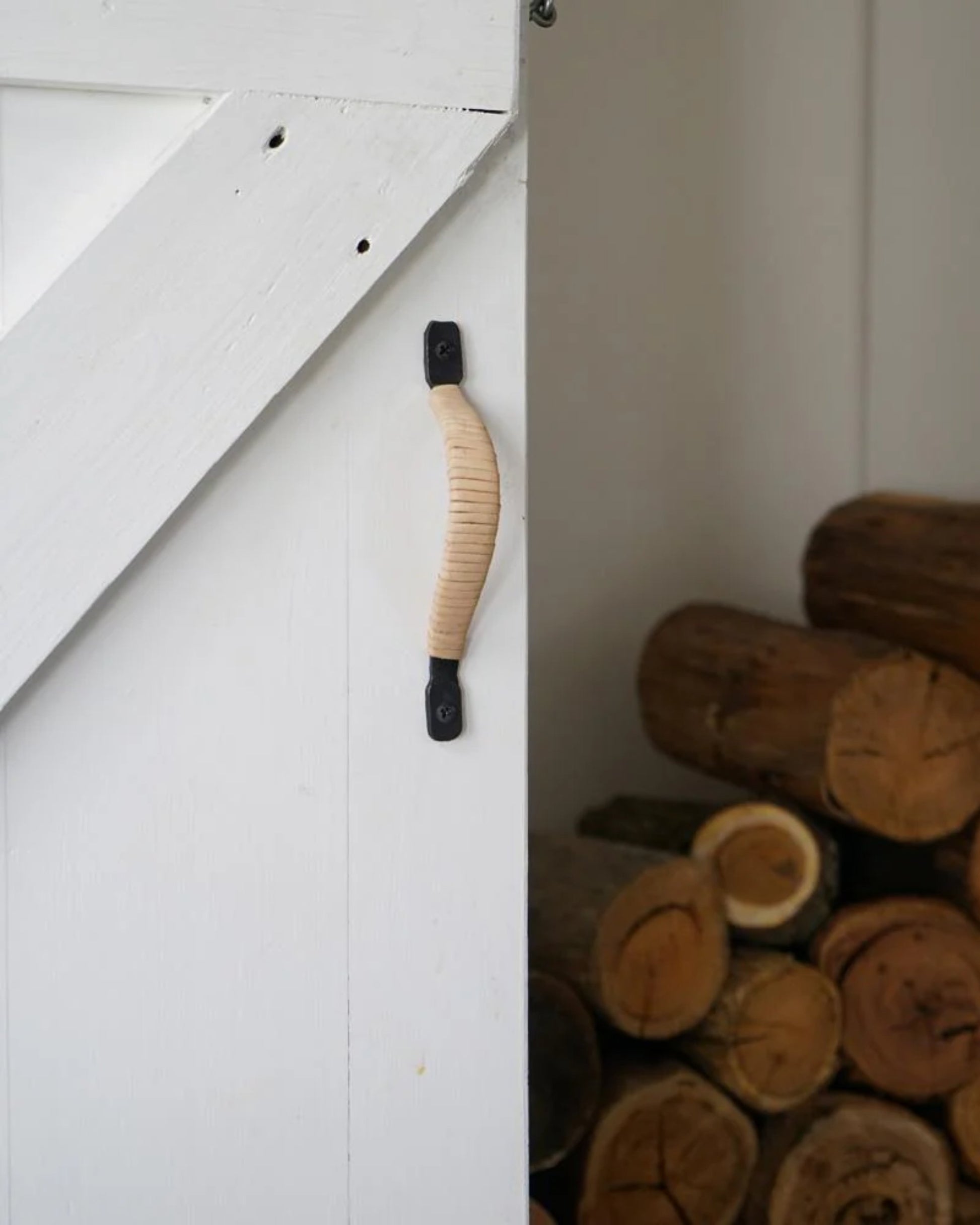 Close-up of a white wooden door showcasing Society Ink's FALCONER HANDLE in leather-wrapped beige. Behind the door, a stack of neatly piled cut firewood rests against a white wall, creating a rustic and cozy atmosphere.