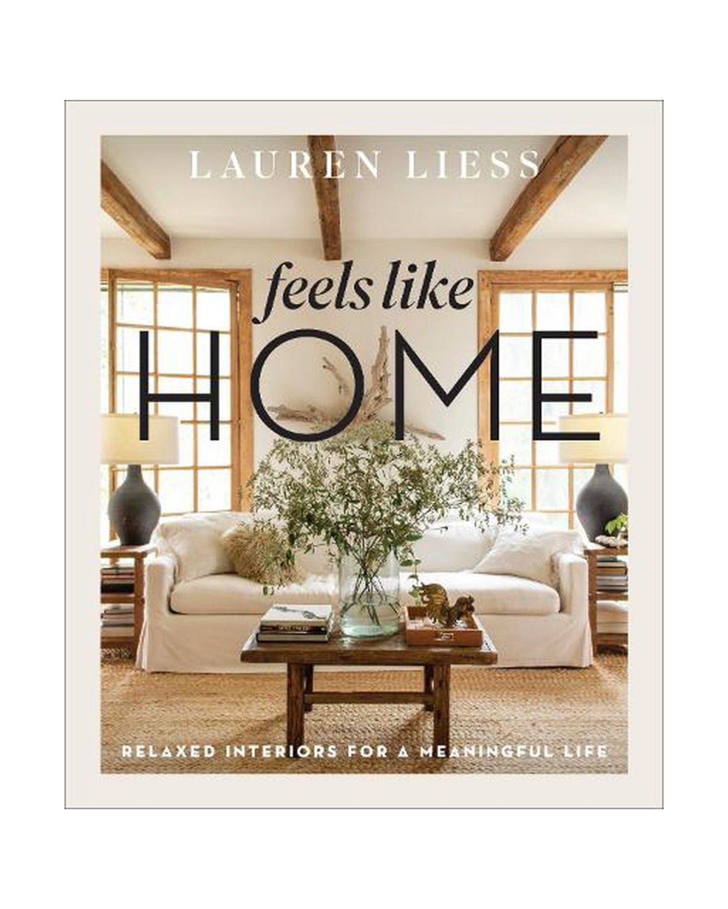 Book cover of "FEELS LIKE HOME: RELAXED INTERIORS FOR A MEANINGFUL LIFE" by Lauren Liess, presented by Bookreps. This design book showcases a cozy living room with a white sofa, wooden beams, large windows, and a coffee table decorated with plants. The tagline reads: Relaxed Interiors for a Meaningful Life and Emotional Connection.